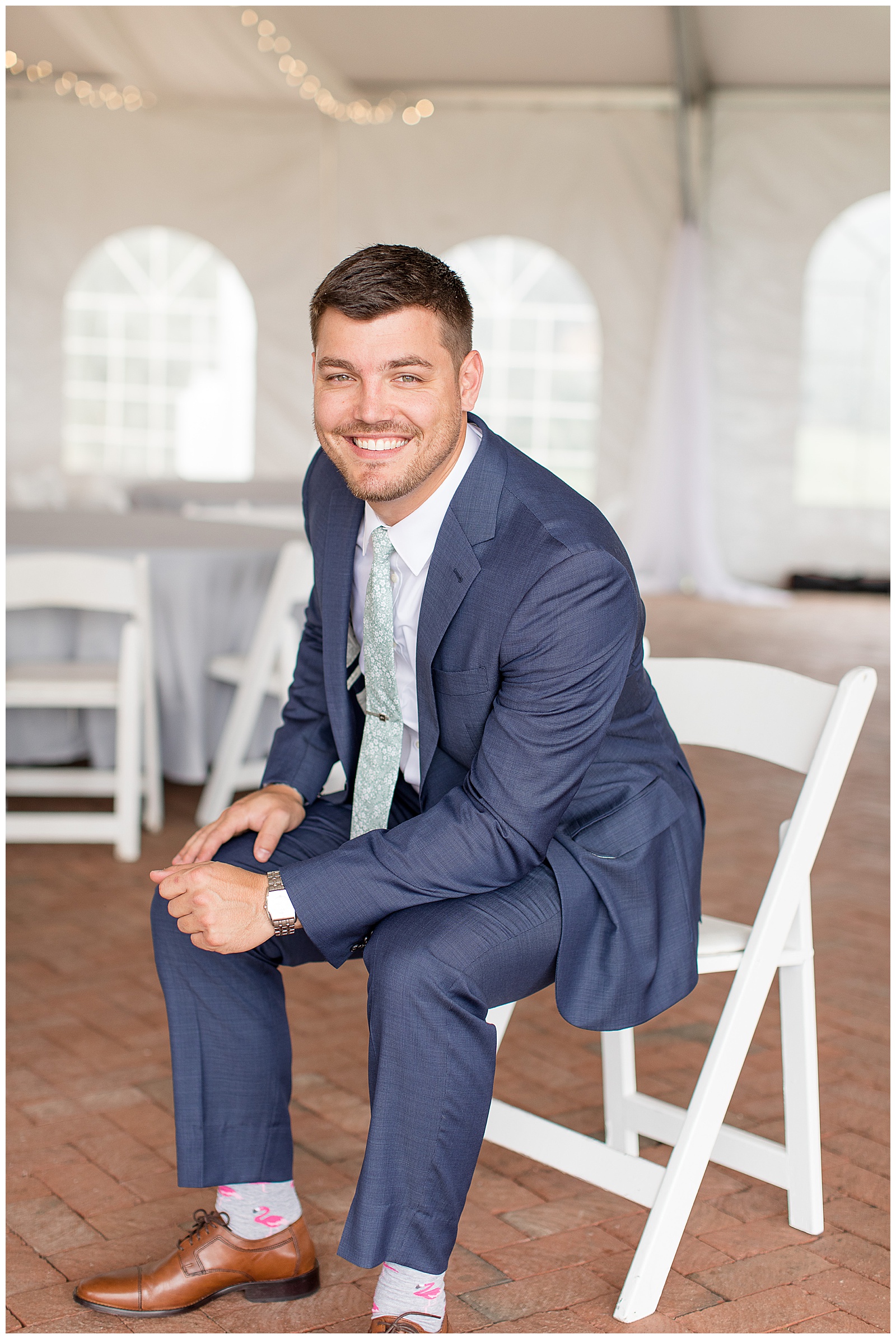 groom sitting in white chair and leaning in towards camera smiling inside white tent at lauxmont farms