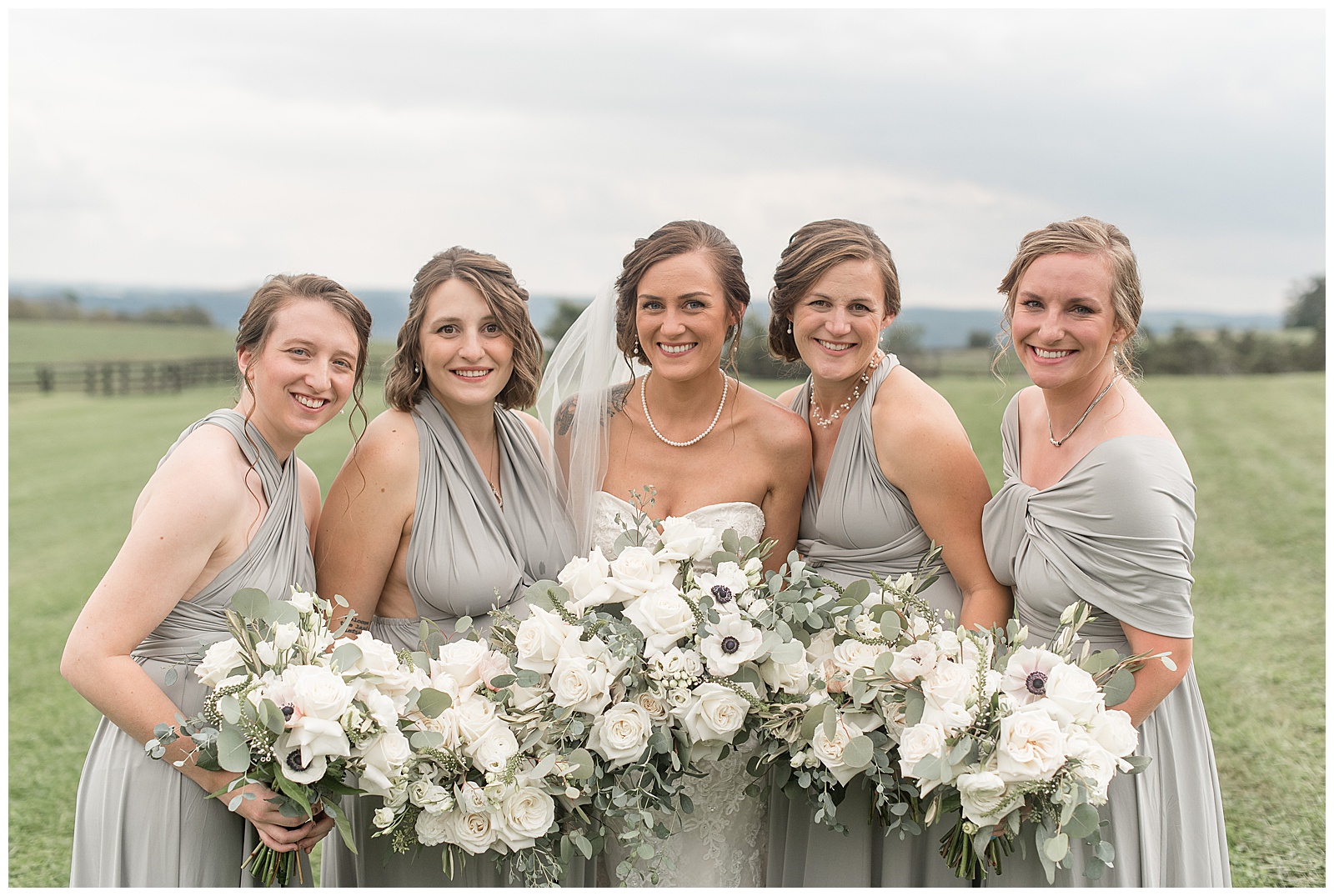 bride surrounded by her four bridesmaids huddled together with their white floral bouquets in front of them in grass field at lauxmont farms