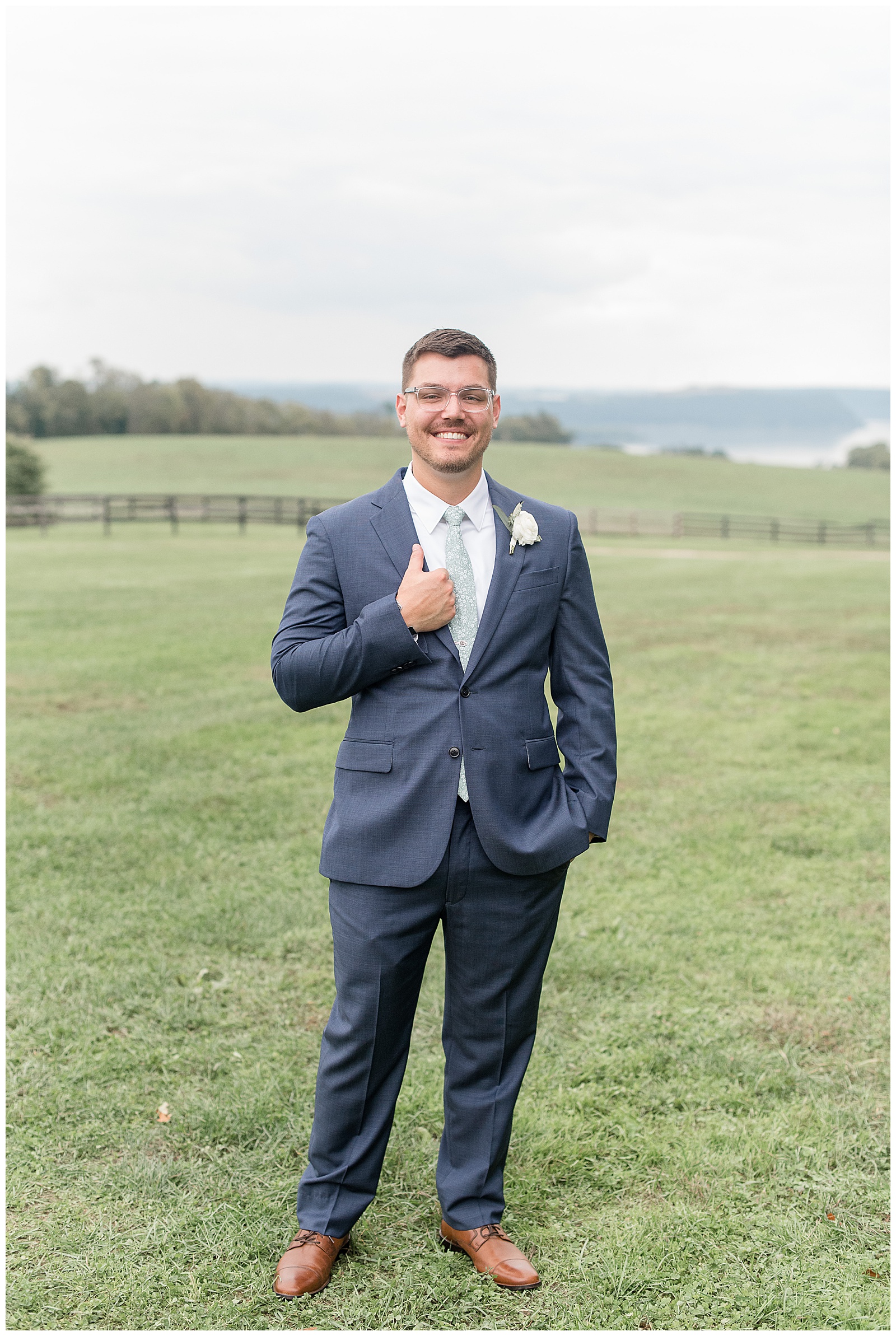 groom holding the edge of his navy blue suit coat and smiling standing out in wide open grass field at lauxmont farms