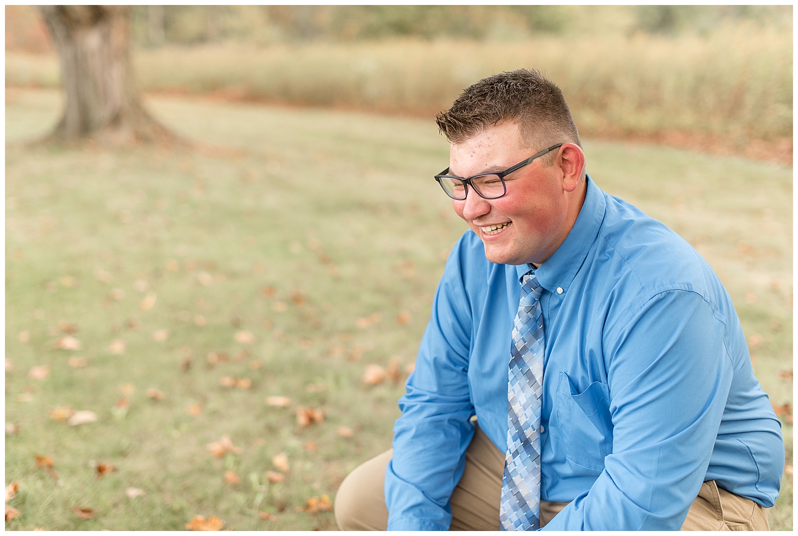 close up photo of senior guy crouching down not looking at the camera and smiling in the grass by field
