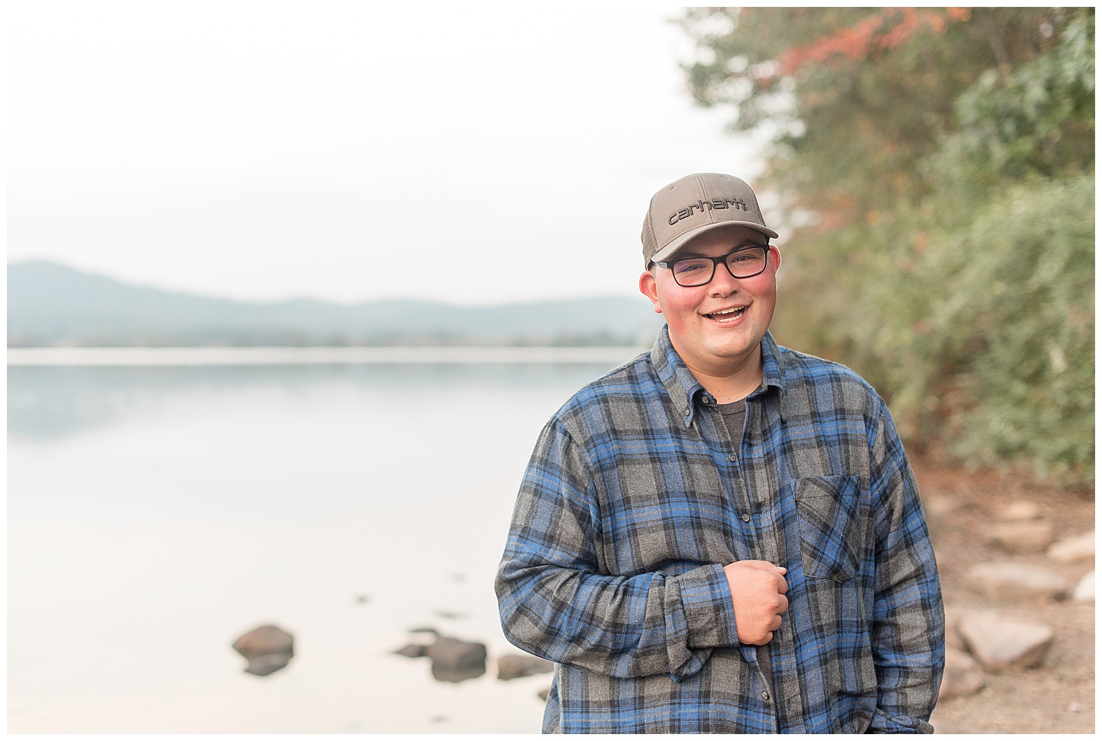 senior guy holding onto middle of his flannel shirt with his right hand smiling with body of water behind him on fall day at middle creek wildlife