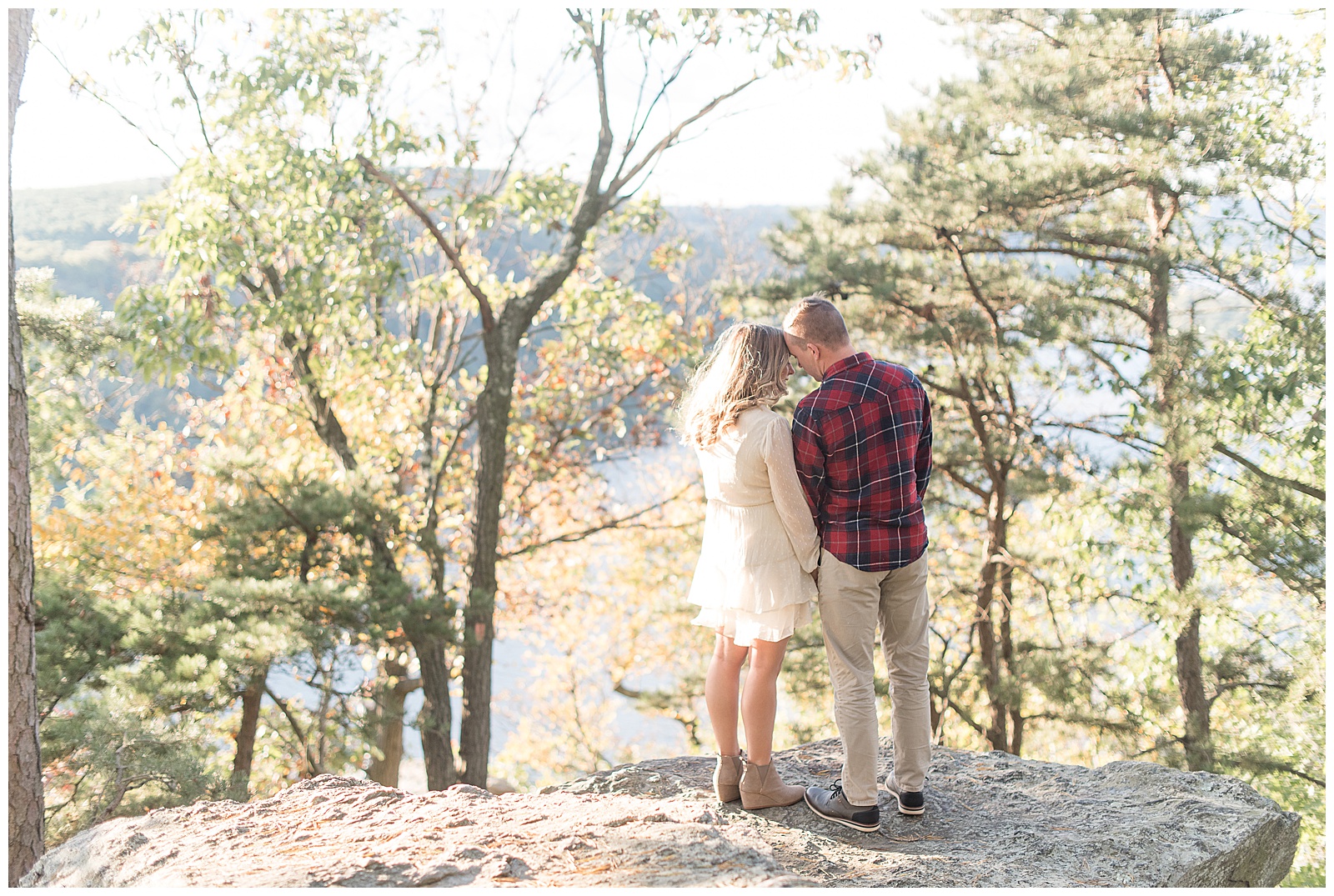 engaged couple standing on large rock with their backs towards camera and their heads touching as the sun shines on them in holtwood pennsylvania