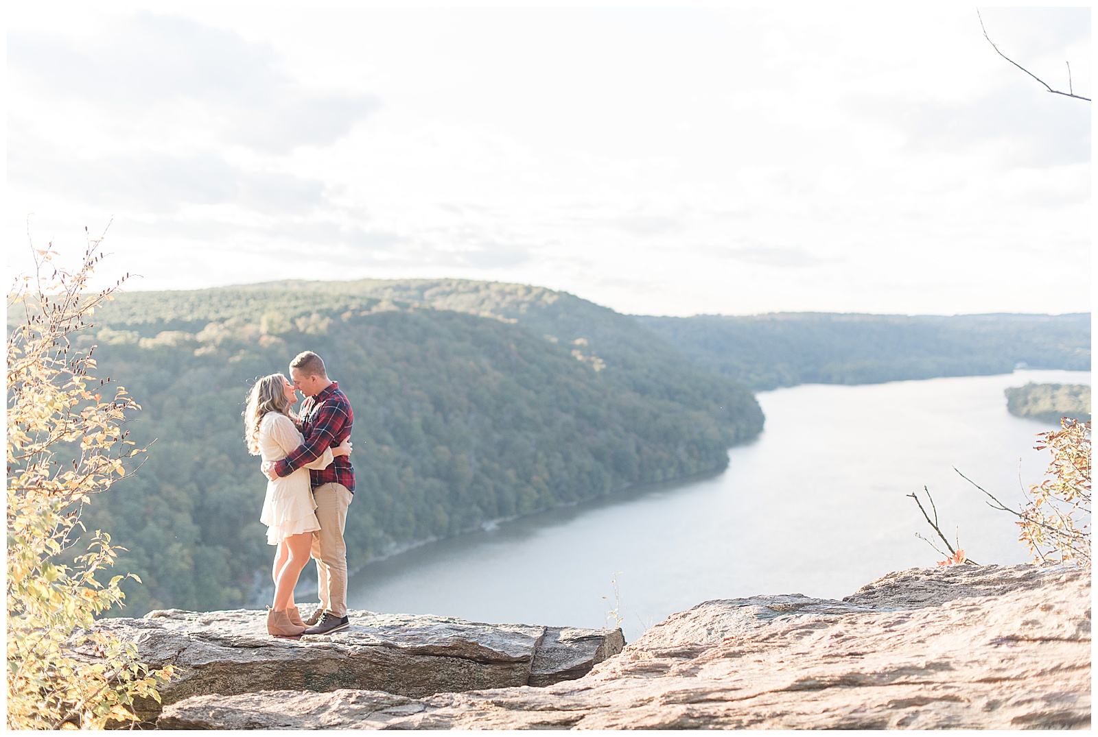 engaged couple hugging and kissing atop large rock at pinnacle point with susquehanna river and hills below them in background in holtwood pennsylvania