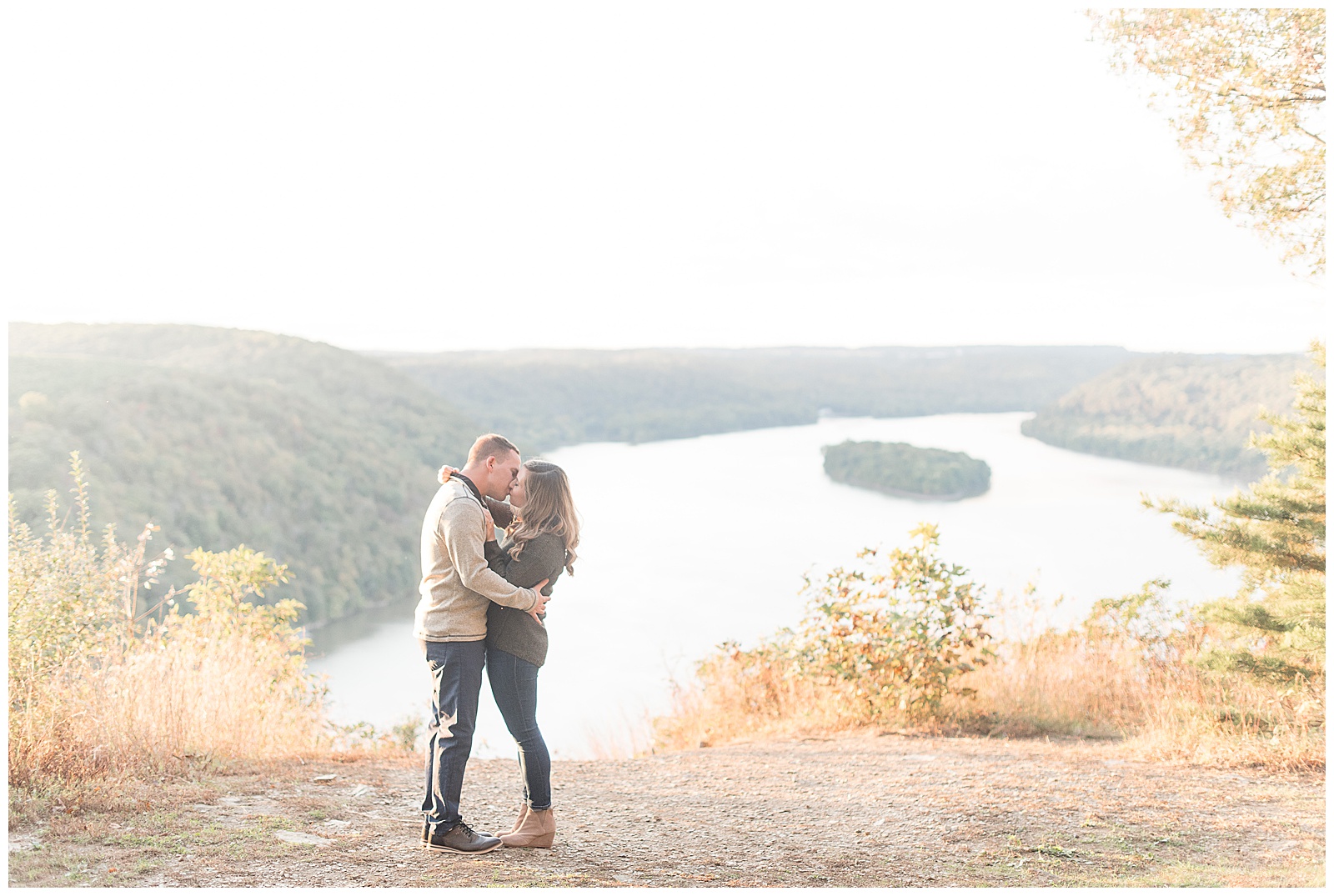 couple hugging and kissing atop pinnacle point with colorful fall leaving and river behind them in lancaster county pennsylvania