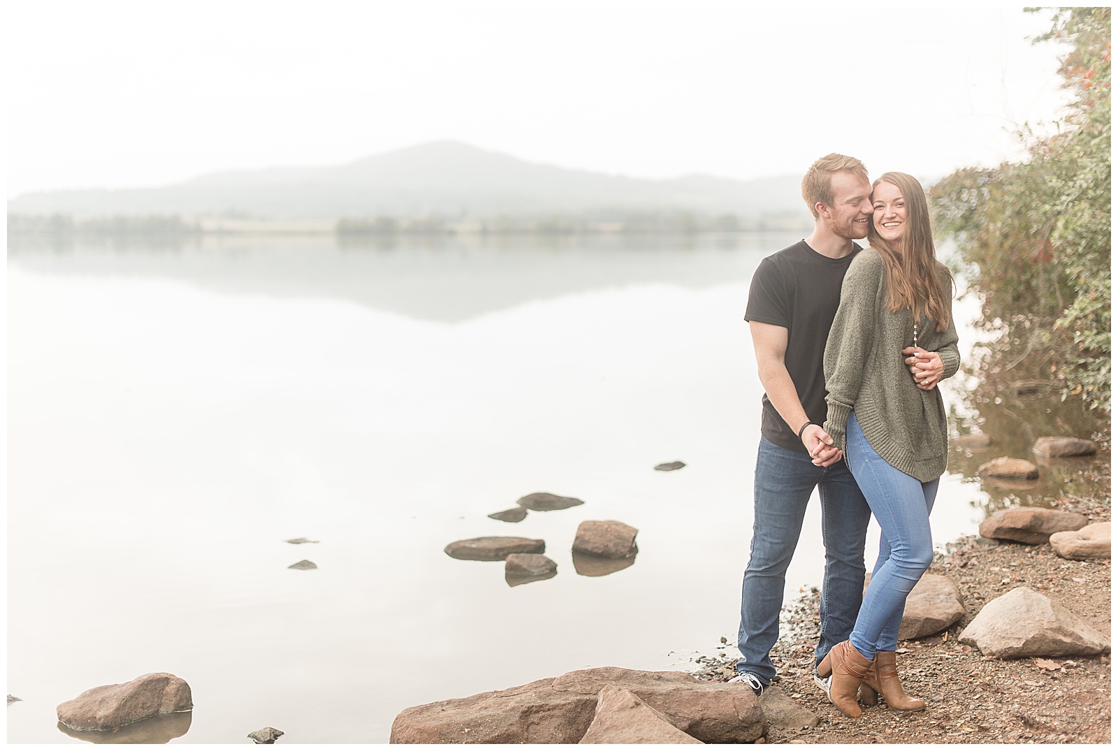 guy hugging girl from behind as they hold right hands and smile with misty lake behind them on cloudy day in lancaster pennsylvania