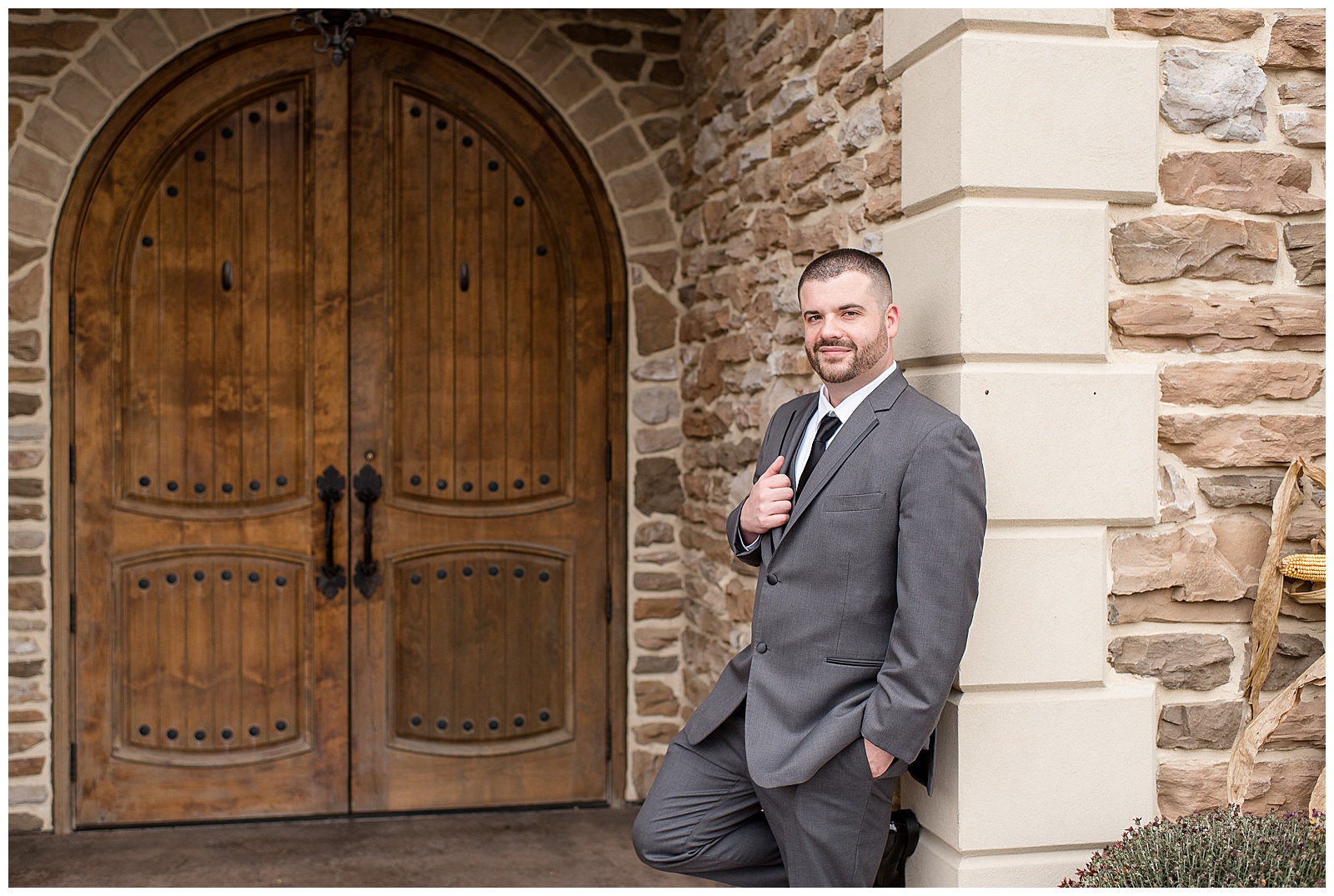 groom leaning against stone building with his right handing holding edge of his suit coat with large wooden door behind him at folino estate