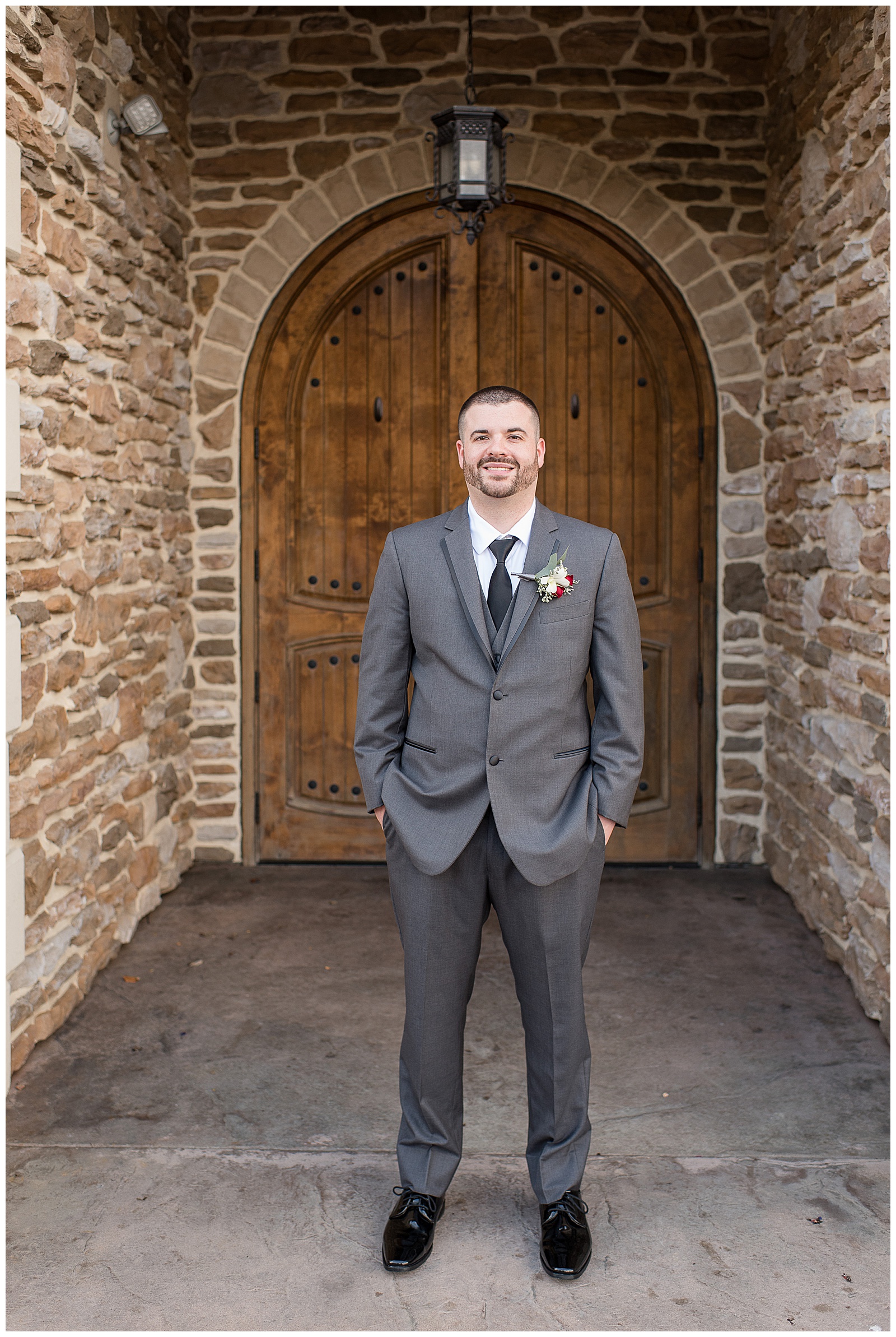 groom standing in front of large wooden door with his hands in his pockets of his dark gray suit and he's smiling at the camera at folino estate