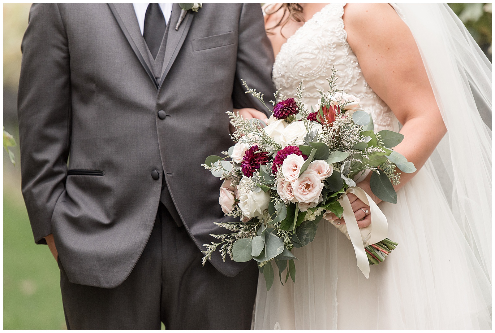 close up photo of groom's dark gray suit coat and bride's gown with her white, light pink, and maroon flower bouquet on display at folino estate