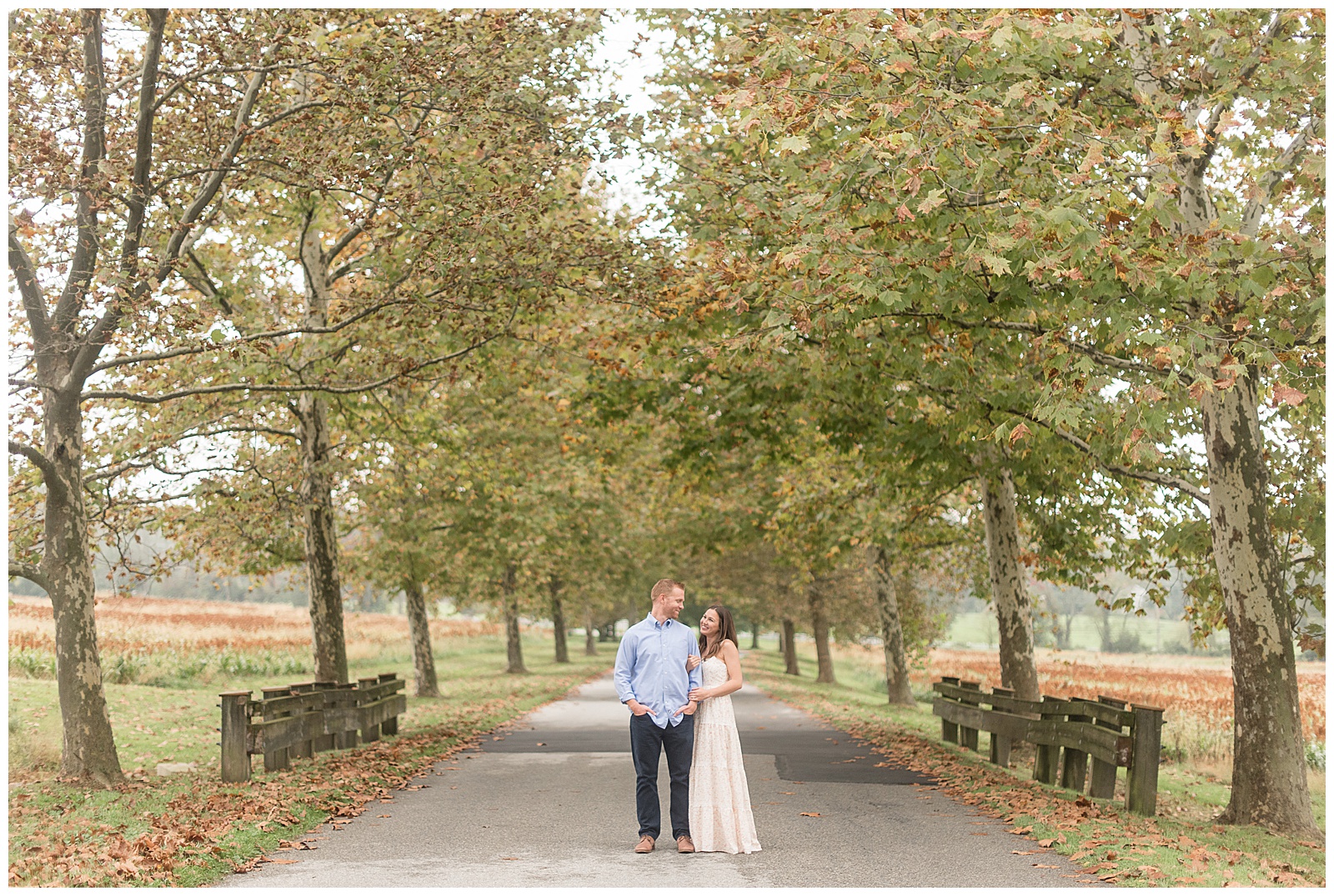 engaged couple standing in middle of tree-lined pathway looking at each other on fall day at springton manor farms