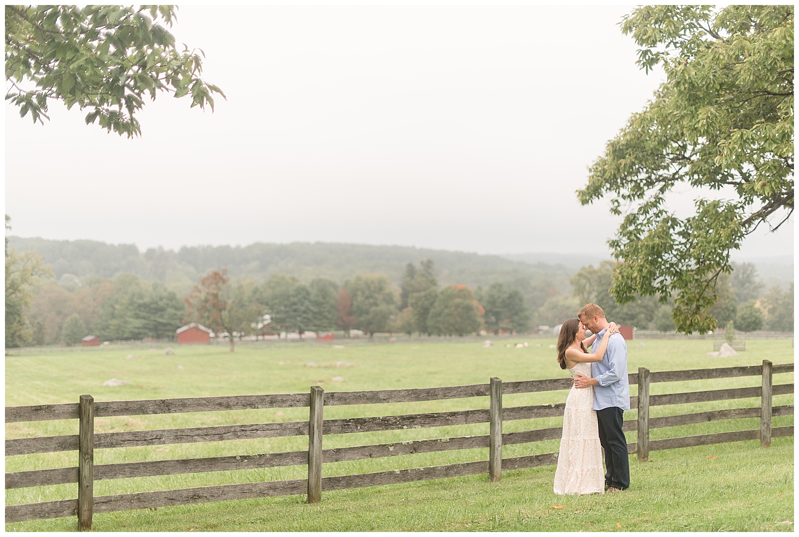 engaged couple hugging tightly on right side of photo with fence and meadow behind them on cloudy day in chester county pennsylvania