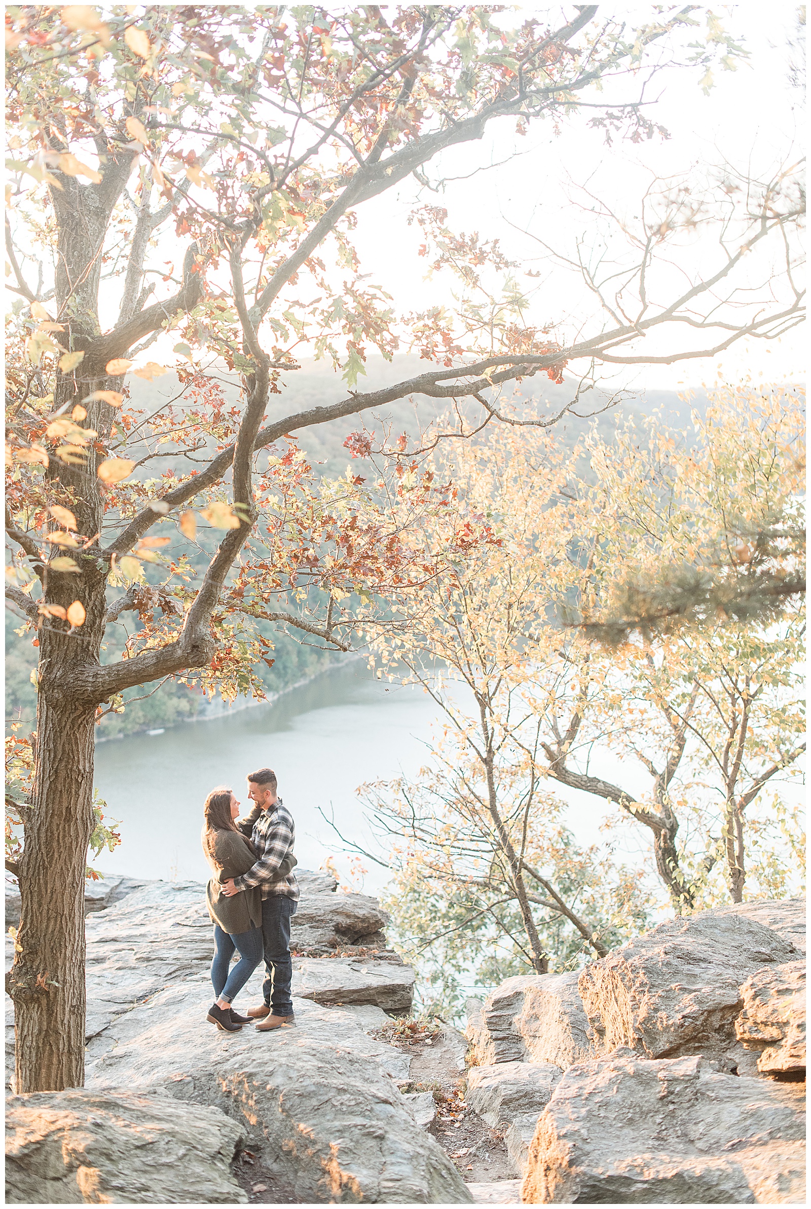 engaged couple hugging and smiling at each other atop large rocks surrounded by colorful fall tress by river at pinnacle point