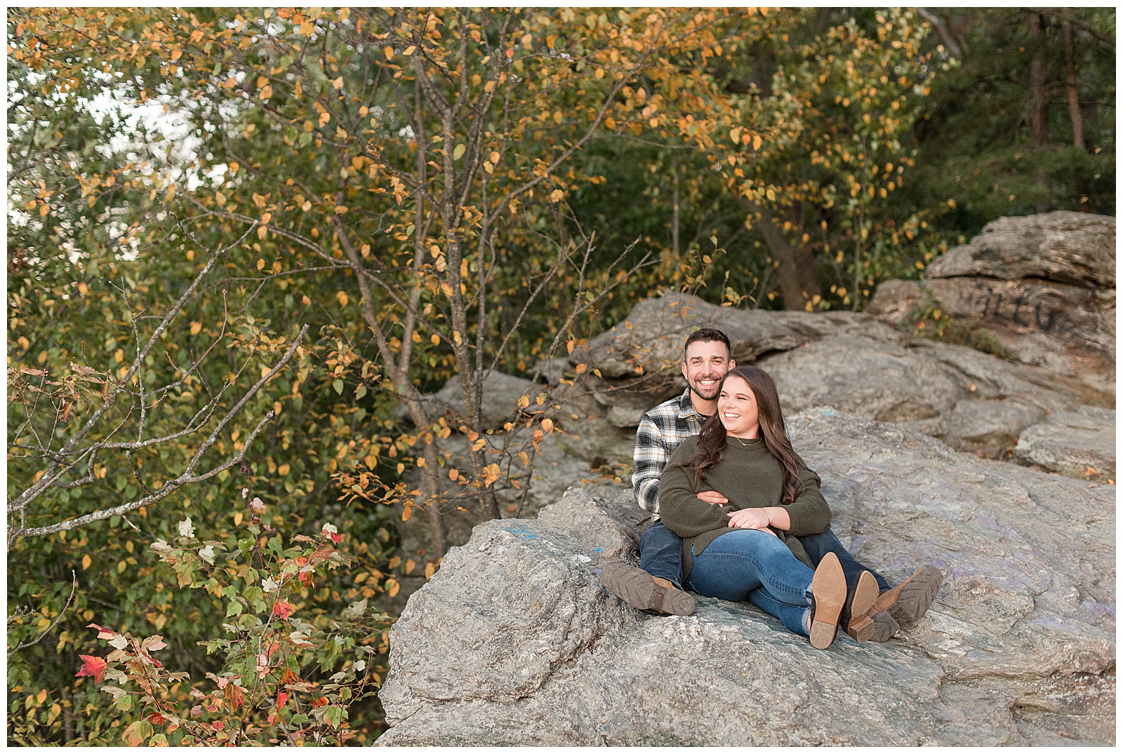 engaged couple sitting atop large rock with their legs extended toward camera and they look towards the right with colorful leaves behind them at pinnacle point