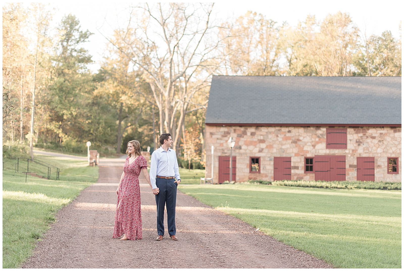 engaged couple holding hands and looking in opposite directions while standing on dirt pathway with historical stone barn behind them in lancaster pennsylvania