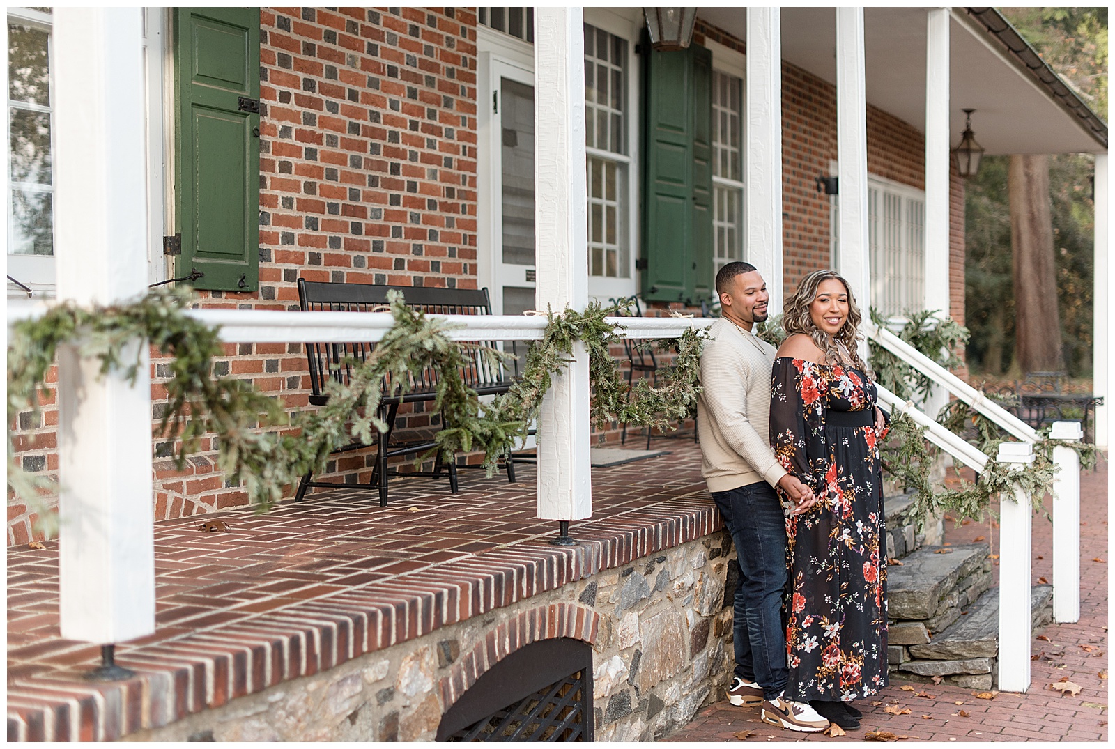 engaged couple standing by brick home's front porch with guy behind girl on fall day at longwood gardens