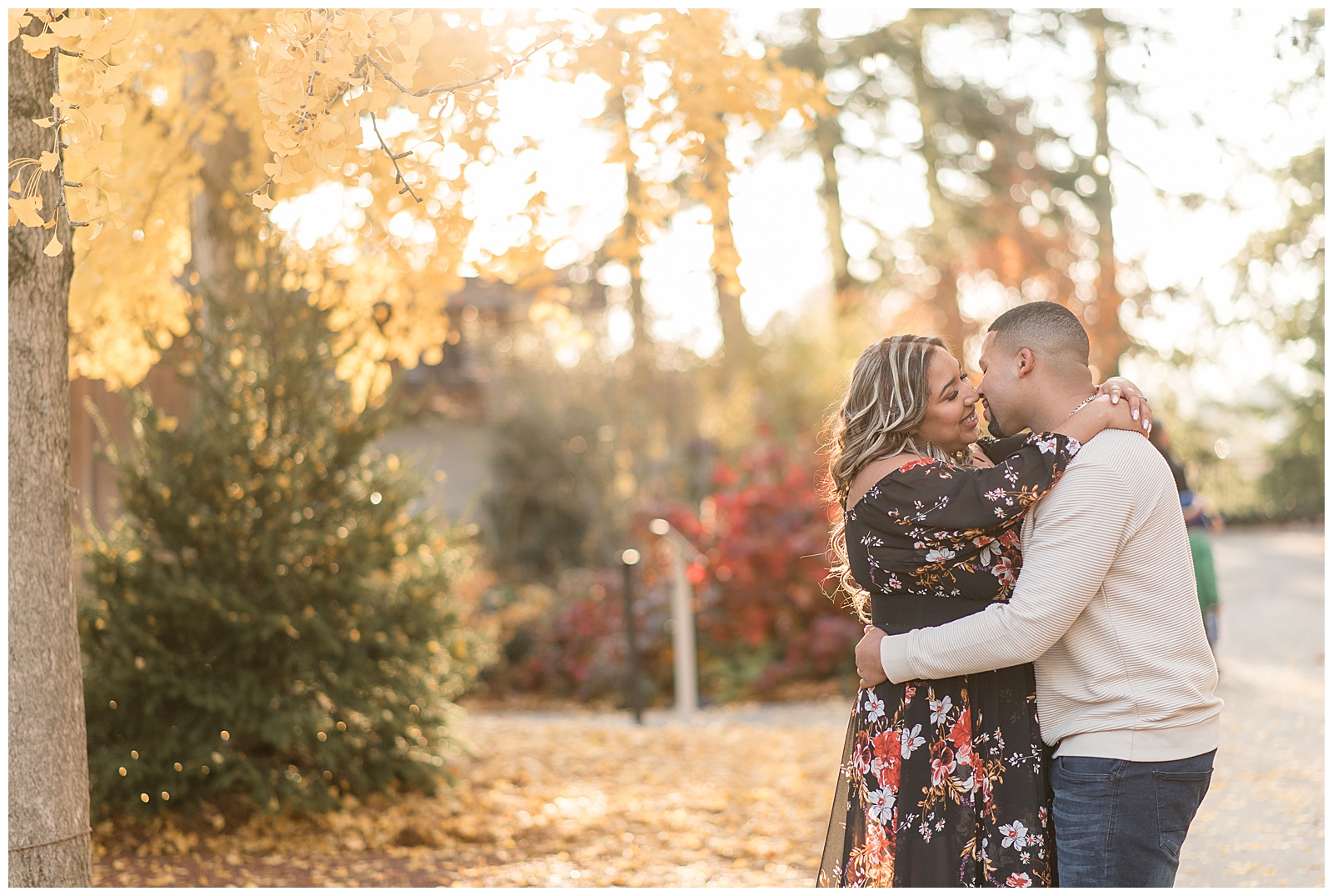 engaged couple hugging and almost kissing on sunny fall day with colorful trees behind them at longwood gardens