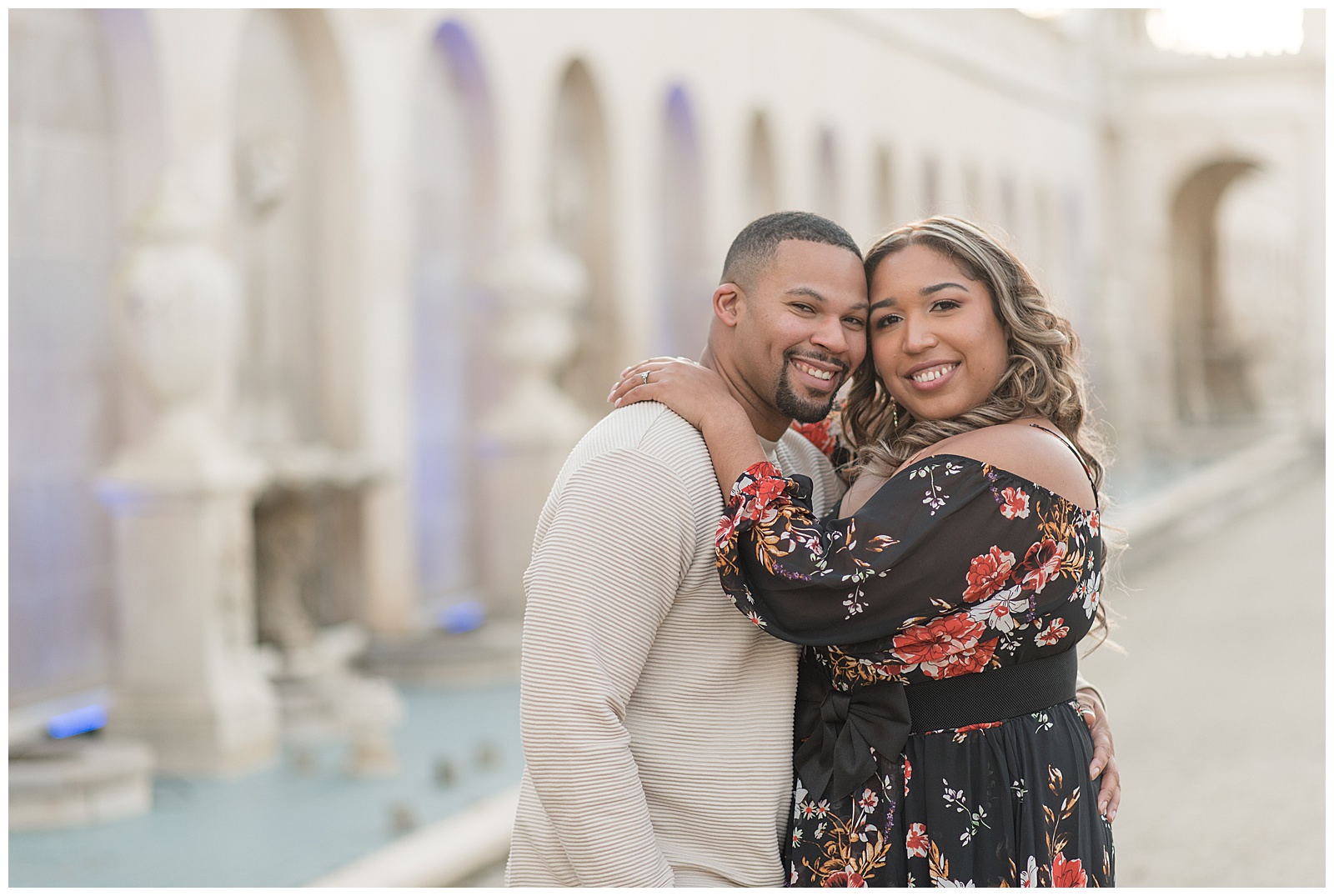 engaged couple smiling at camera as woman wraps her arms around guy's shoulders with concrete archways and fountains behind them at longwood gardens