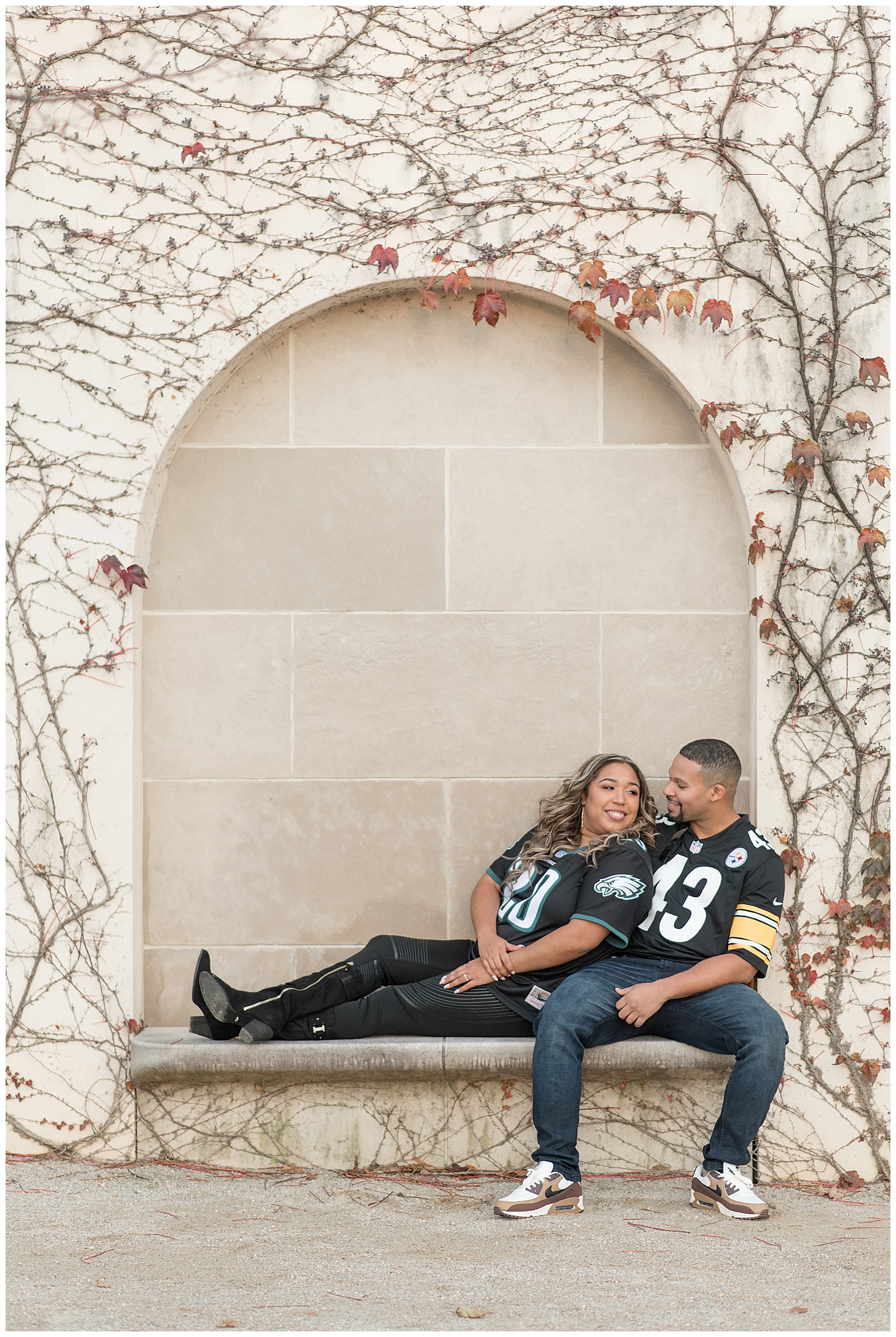 engaged couple wearing pittsburgh steelers jerseys and jeans sitting on concrete bench with woman extending her legs out on bench at longwood gardens