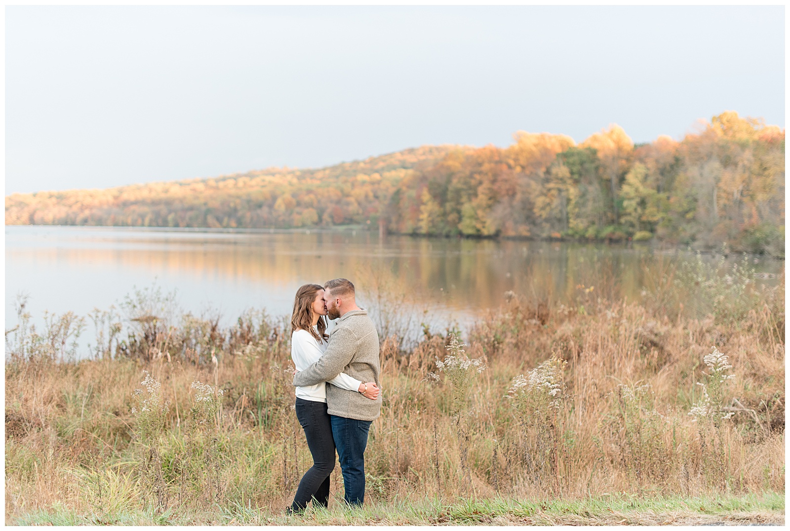 engaged couple hugging as they each are wearing a sweater and dark pants by beautiful lake at middle creek wildlife on fall day