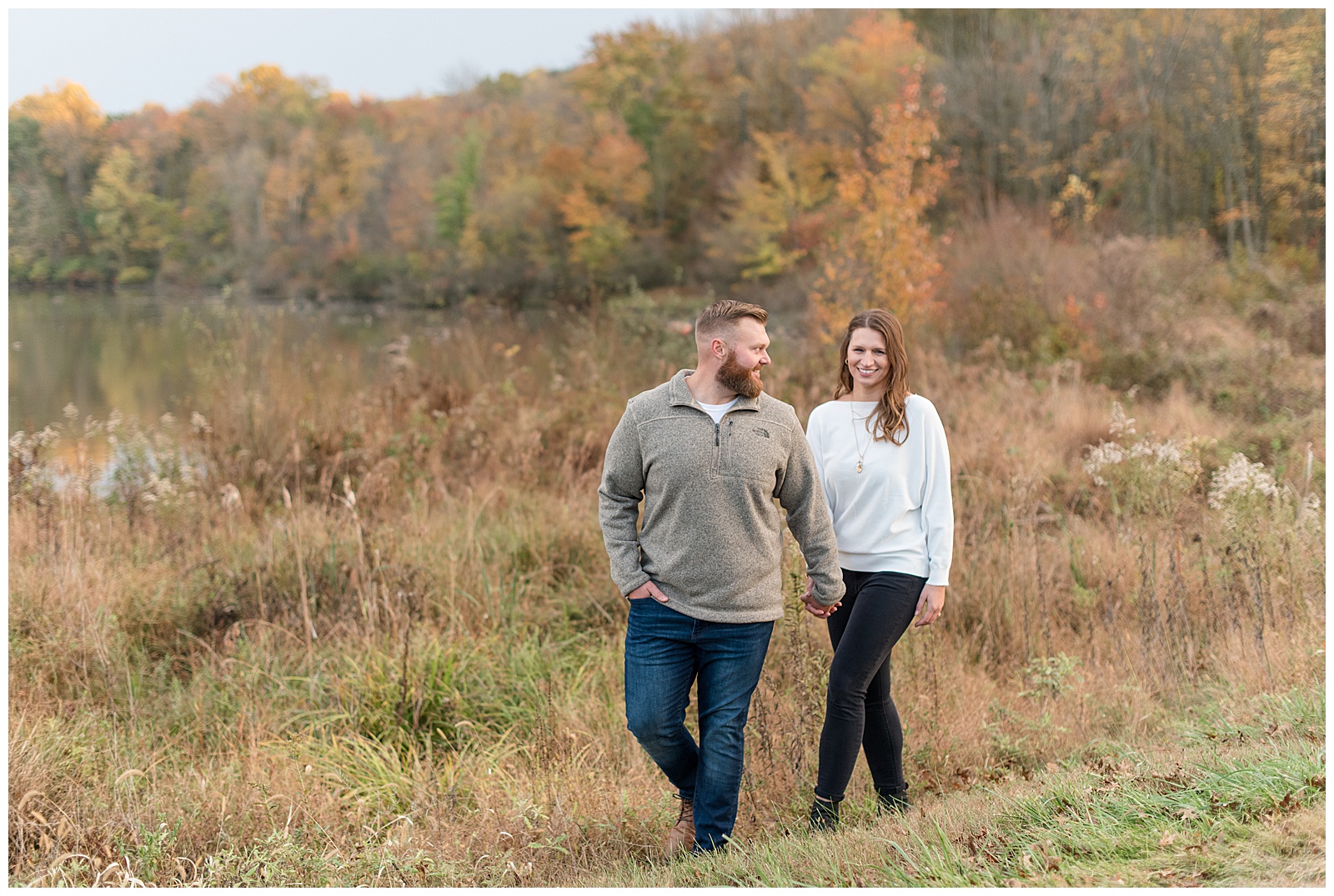 engaged couple holding hands and walking along wild grasses on colorful fall day in lititz pennsylvania