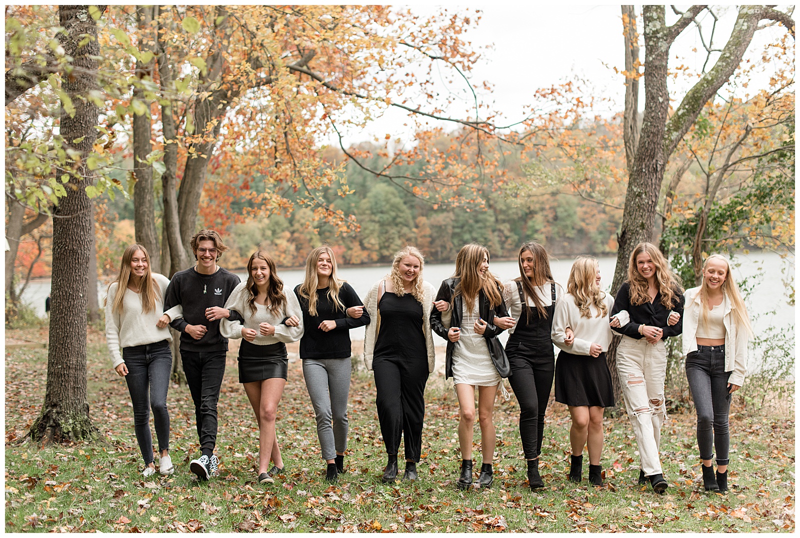 nine senior girls and one senior guy with arms all linked walking towards camera smiling at reservoir park on fall day in york pennsylvania