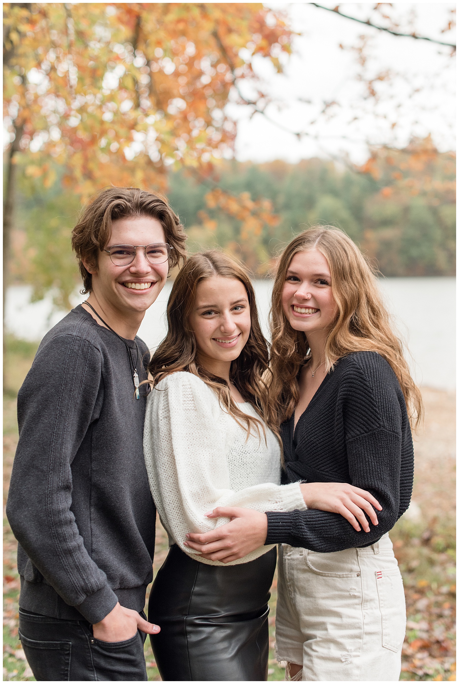 senior guy standing with two senior girls as they all smile with bright orange tree leaves and lake behind them in york pennsylvania