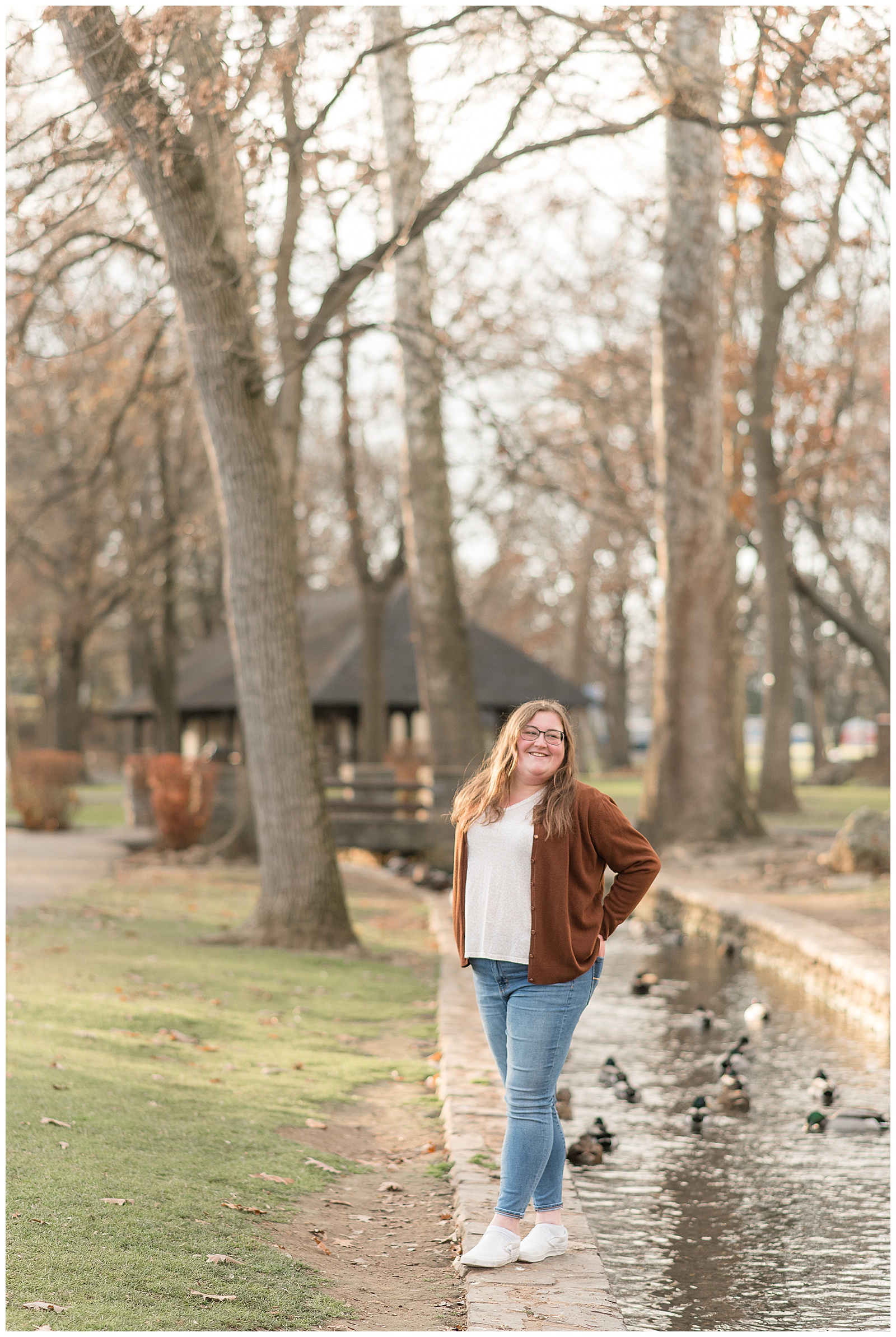 senior girl wearing jeans with brown and white shirt with hands on hips by stream running through lititz springs park with ducks in the water