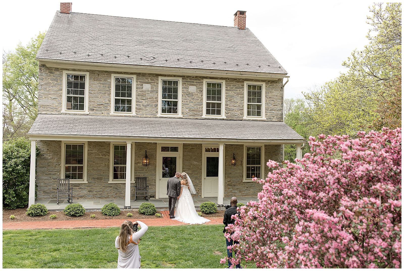 back of photographer as she shoots photos of bride and groom standing by steps to front porch of historic gray stone house in southeastern pennsylvania