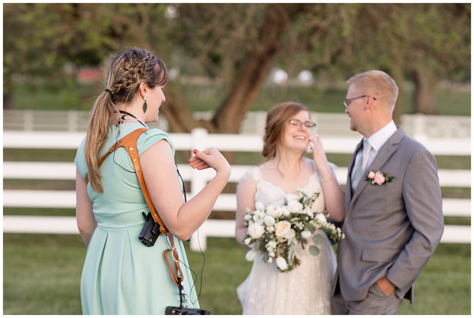 photographer chatting with bride and groom on sunny day by white fence at the barn at silverstone in lancaster pennsylvania