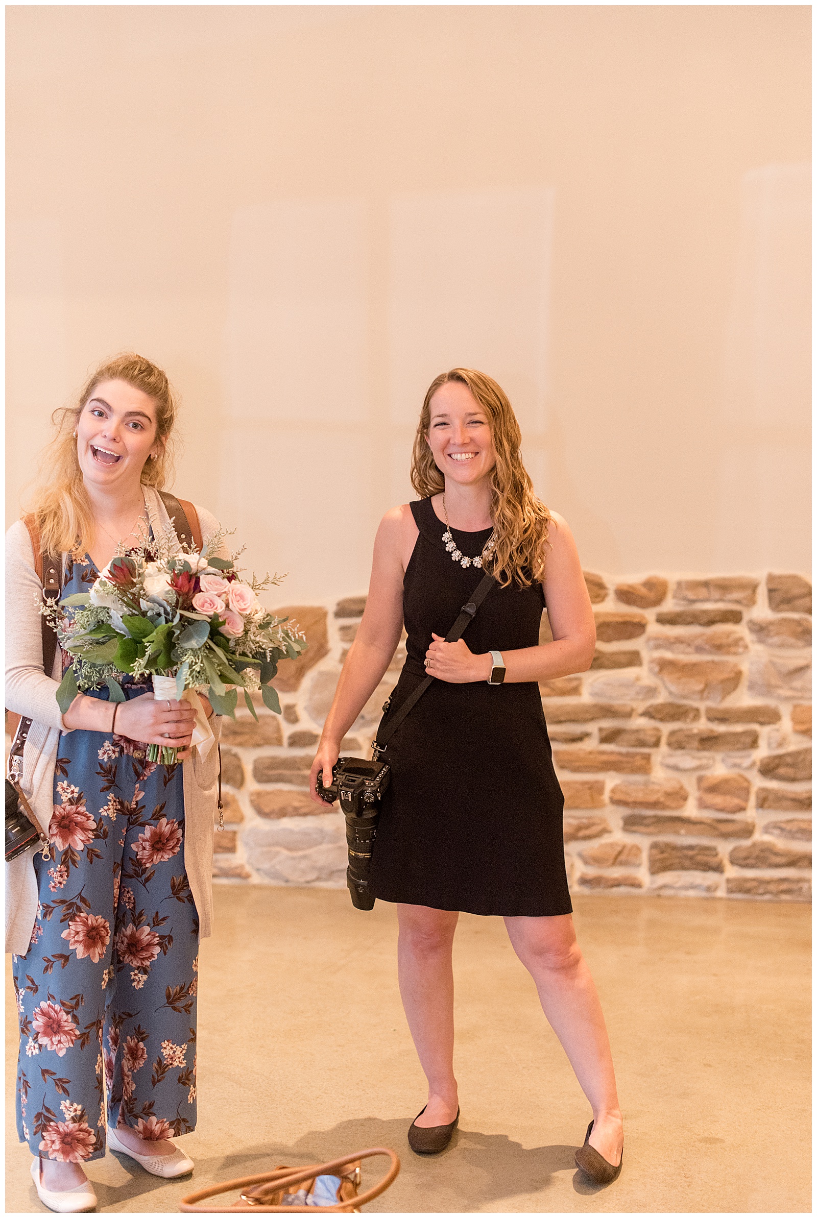 two photographers posing for a silly candid moment as one holds bride's bouquet and the other holds camera inside wedding venue with stone walls