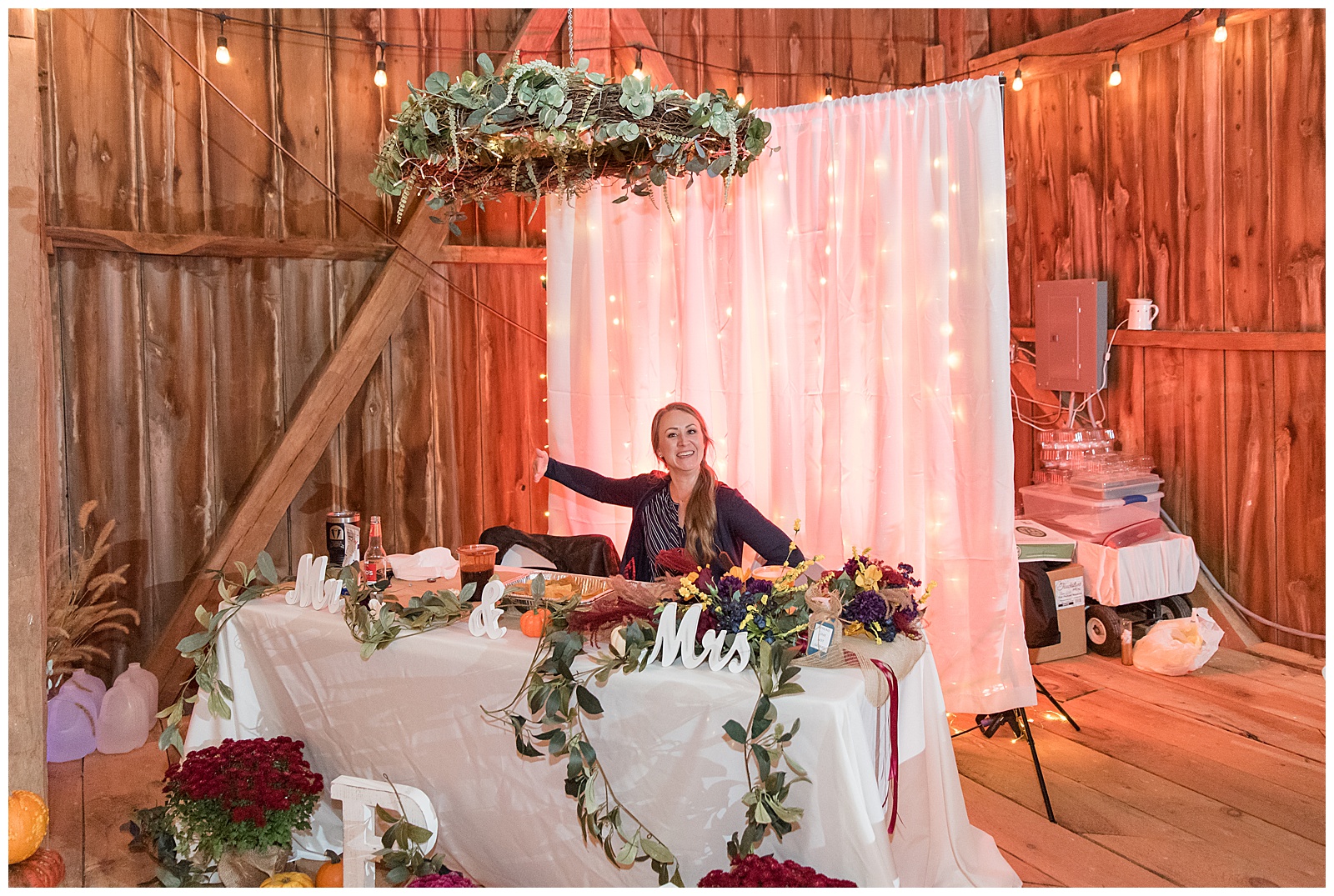 photographer sitting behind table prepared for bride and groom for reception and smiling for camera as they take photos before reception begins with white backdrop and large floral display overhead