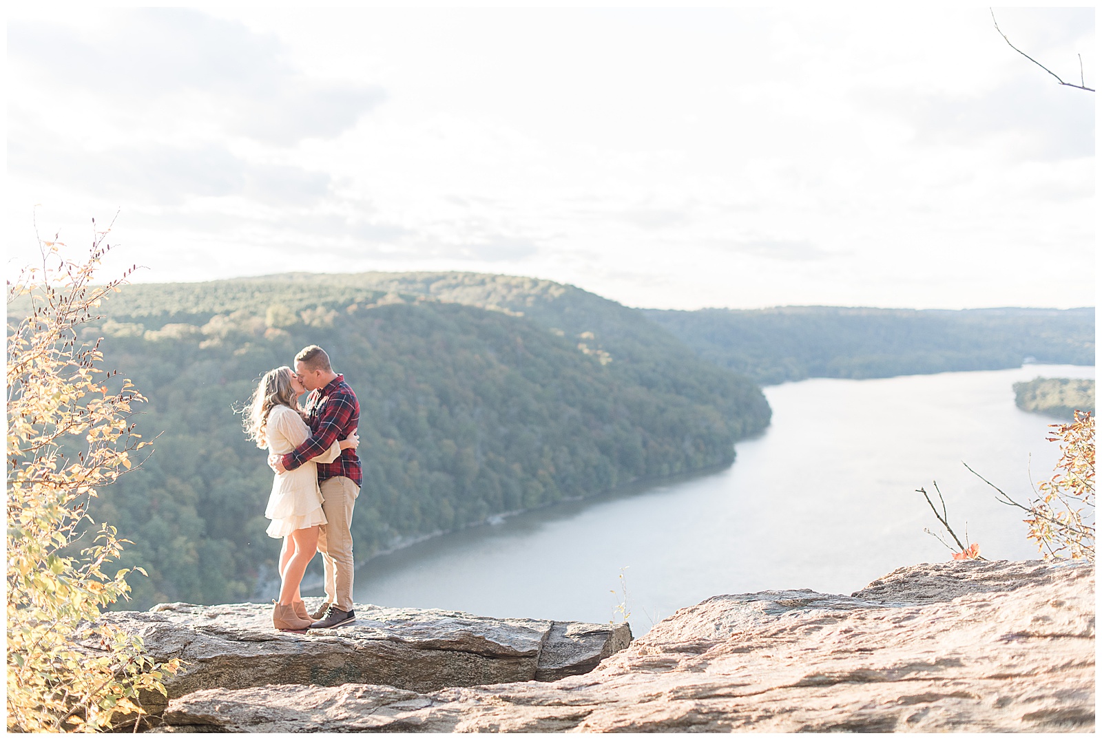 engaged couple kissing with their arms around each other atop large rock at pinnacle point overlook with river below them in background on sunny evening in holtwood pennsylvania