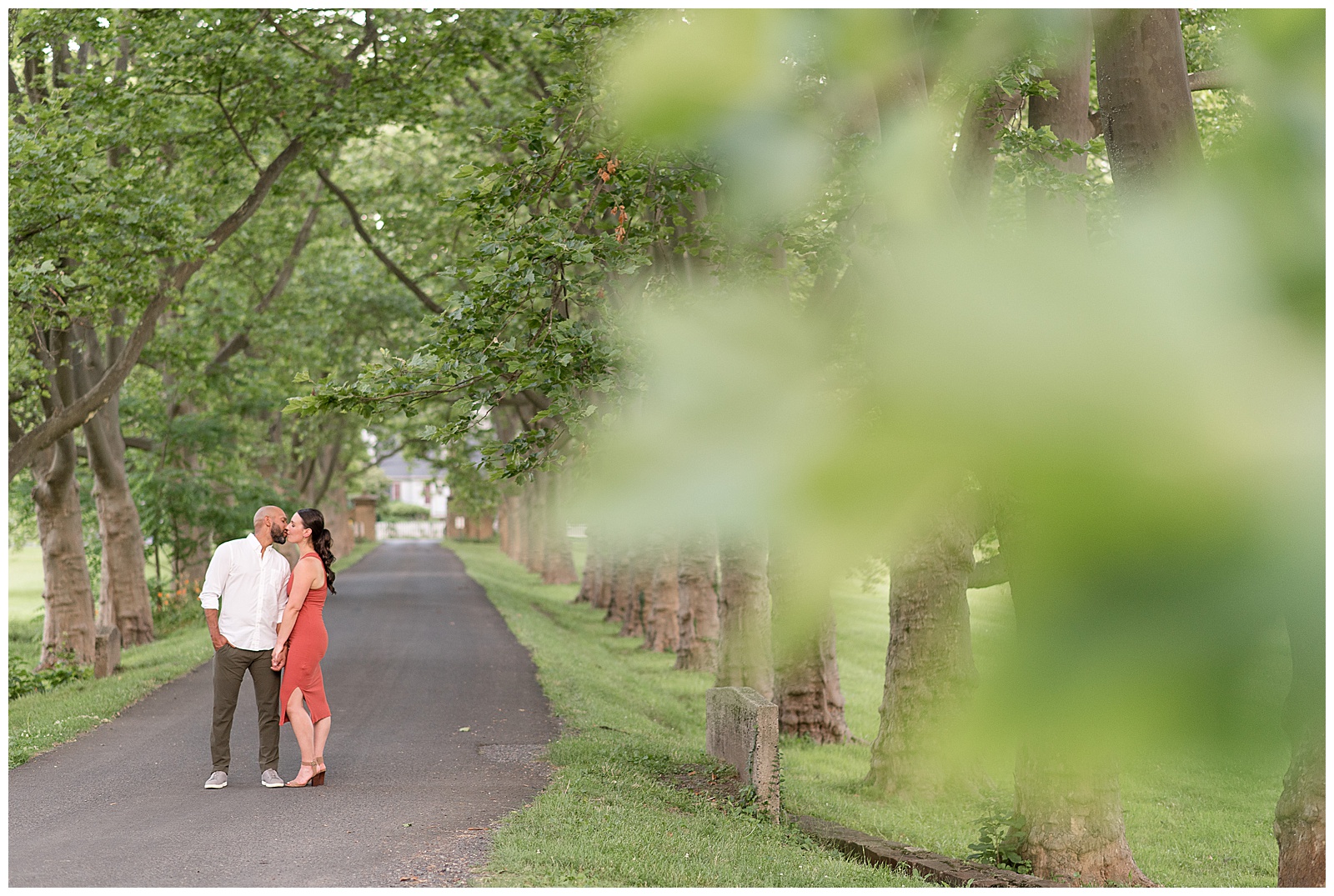 engaged couple standing close and man has right hand in his pocket along paved tree-lined pathway on sunny evening