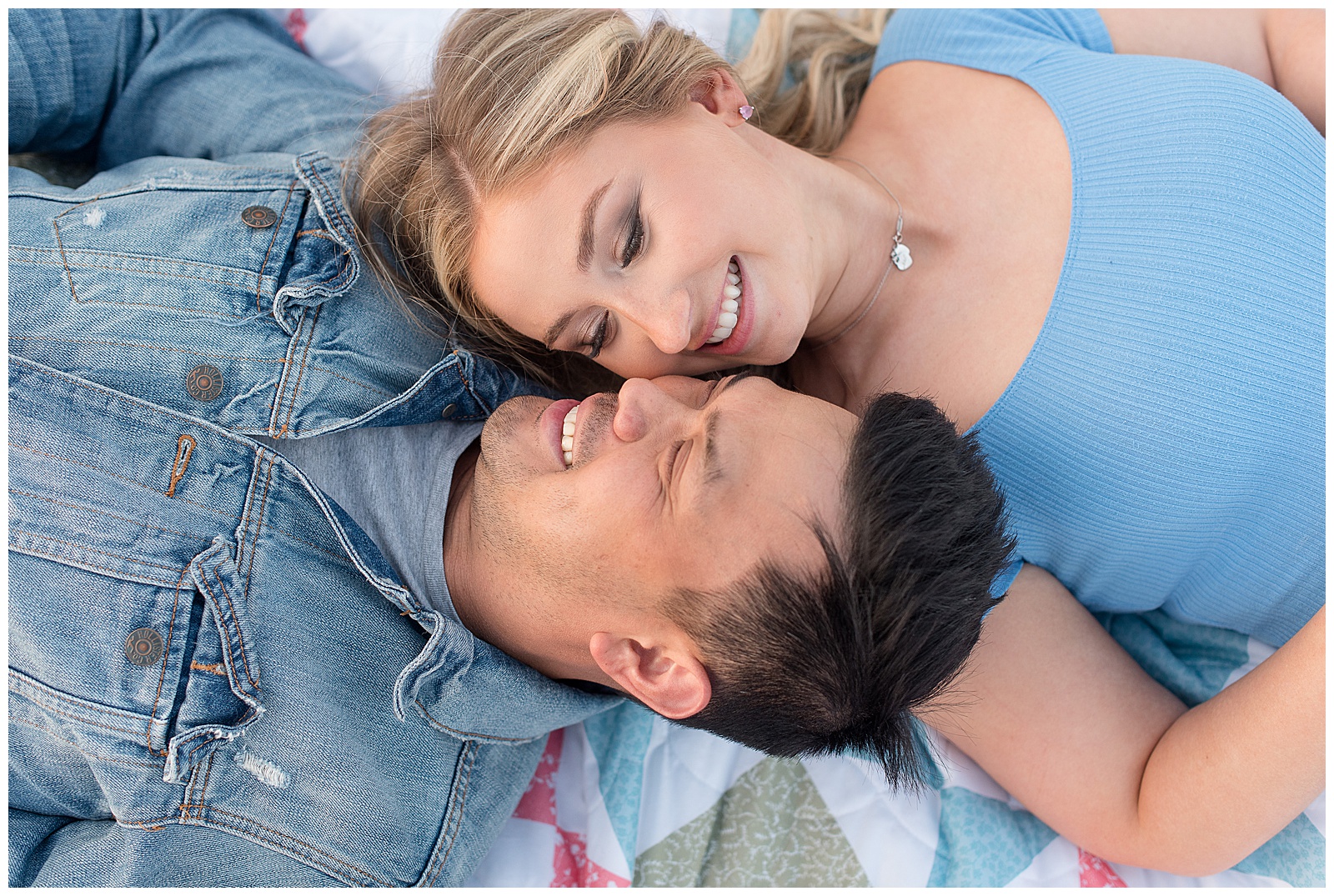 engaged couple laying on the ground on quilt with their faces nestled around one another as they smile and each are wearing blue shirts