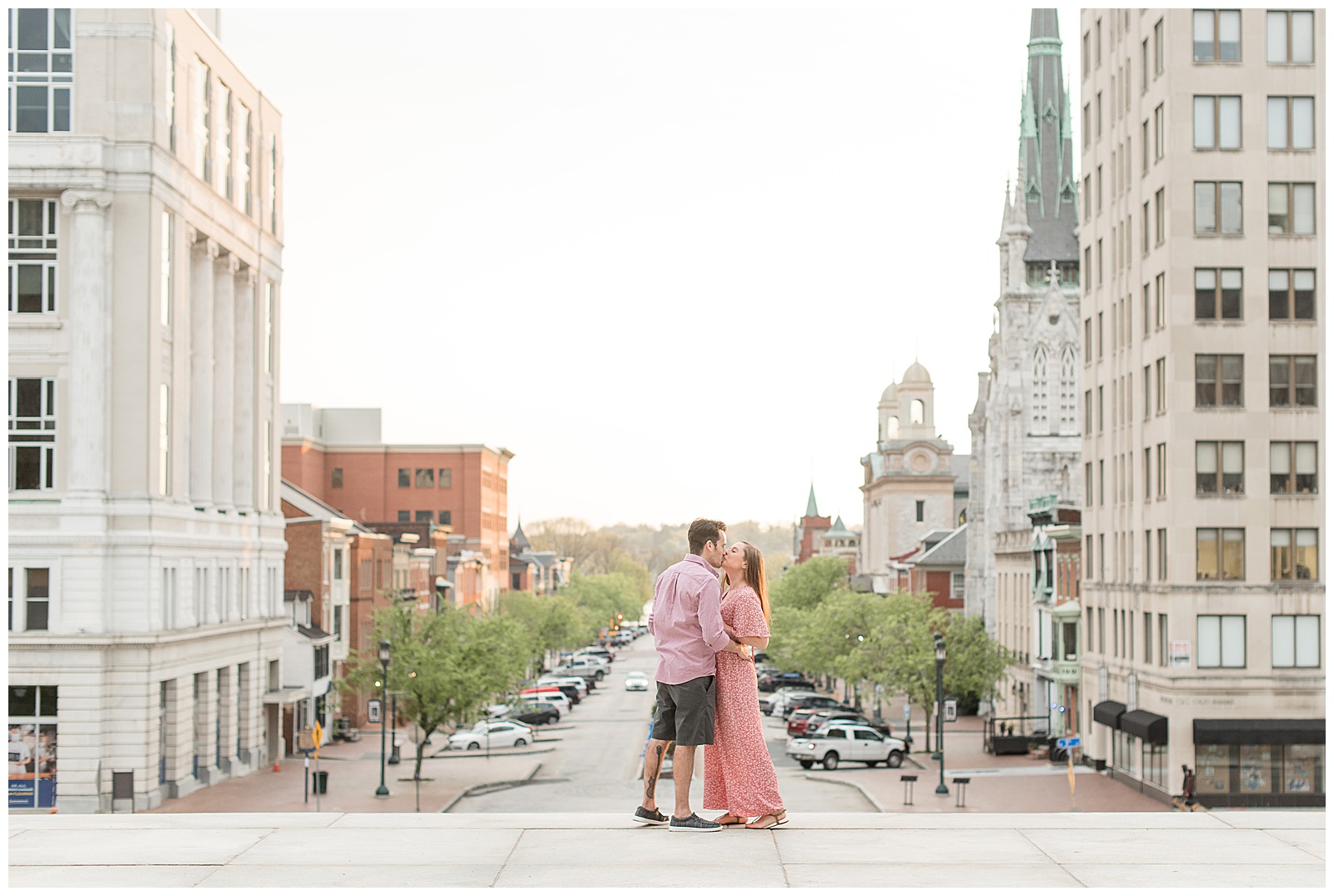 engaged couple standing on sidewalk kissing with city buildings behind and below them on sunny evening in harrisburg pennsylvania