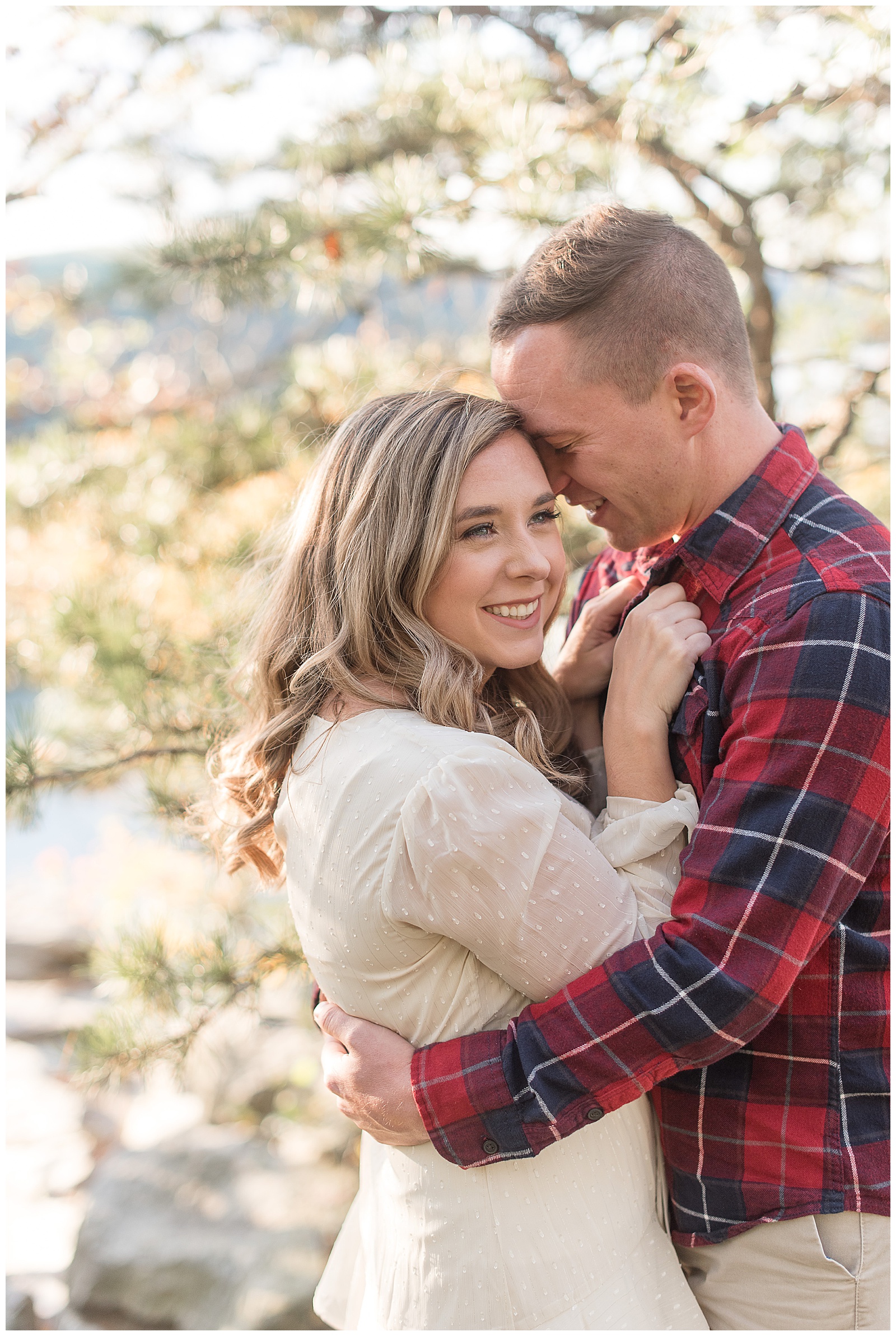 guy wrapping his arms around girl as she holds onto his flannel shirt and looks off into the distance smiling with trees behind them