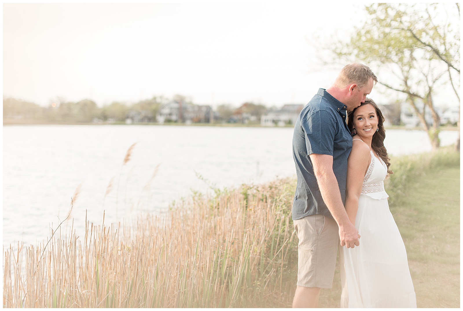 couple standing along bay by wild grasses with guy standing behind girl as they hold hands and he kisses top of her head