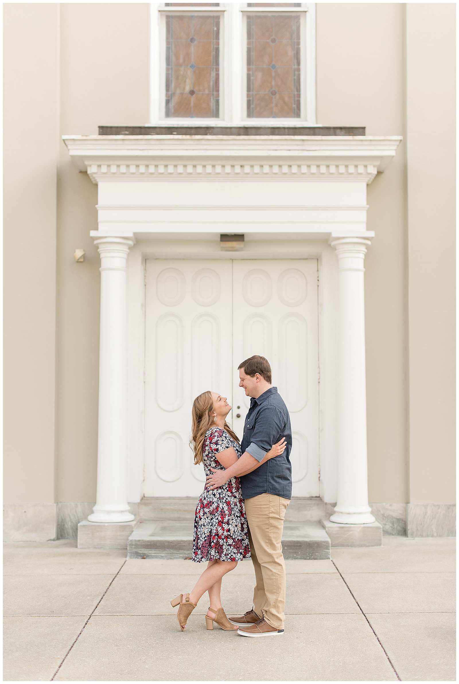engaged couple standing on sidewalk in front of tan home with large white front door with white columns on bright evening as they hug and smile