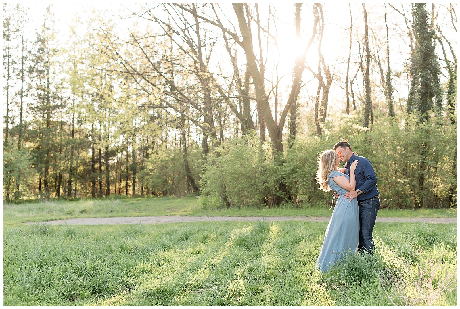 engaged couple kissing on right side of photo with woman leaning back slightly and sunset shining through row of trees at manheim township park in lancaster pennsylvania