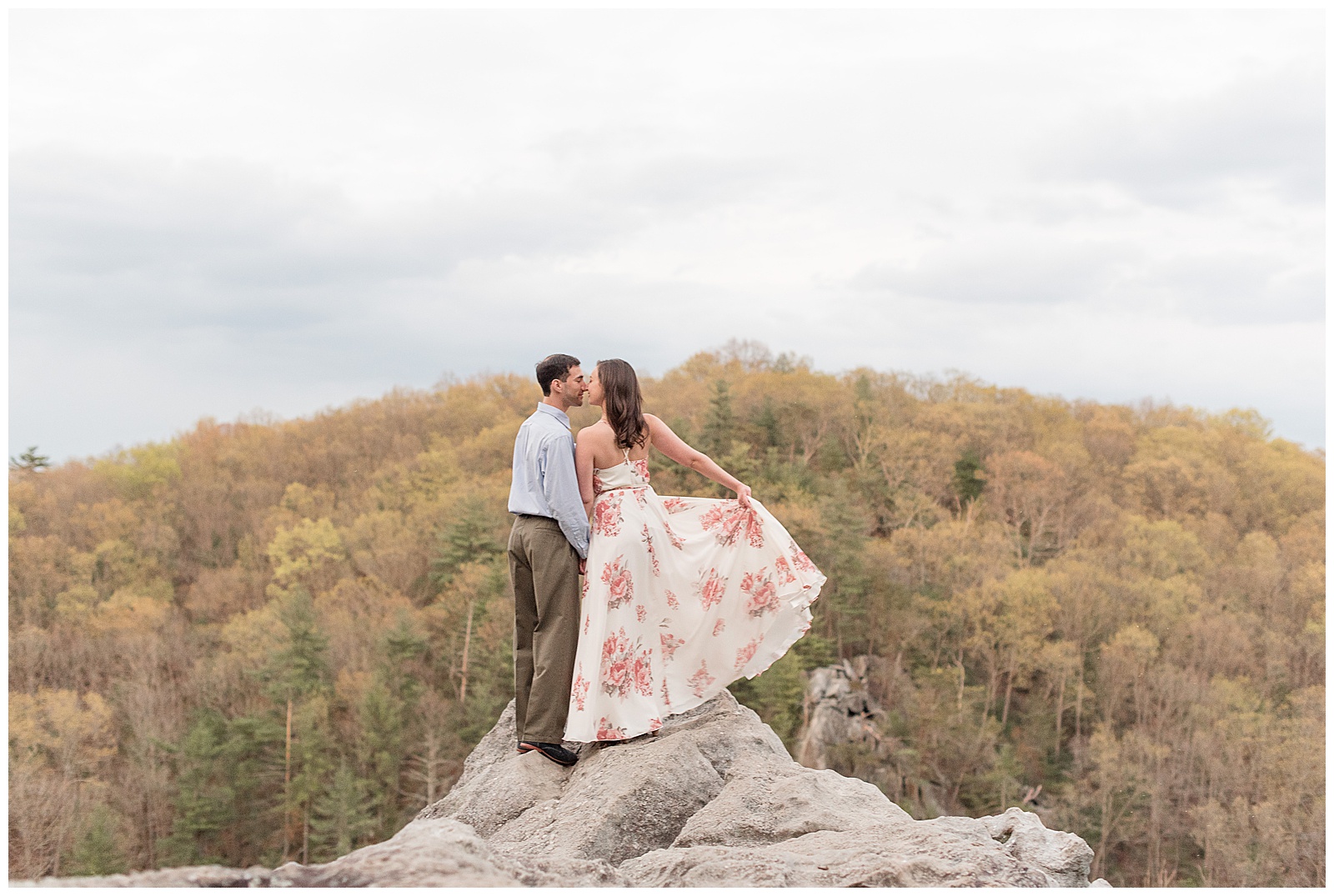 engaged couple standing with backs toward camera atop pointy rock by trees as they kiss and she holds out long dress with her right hand