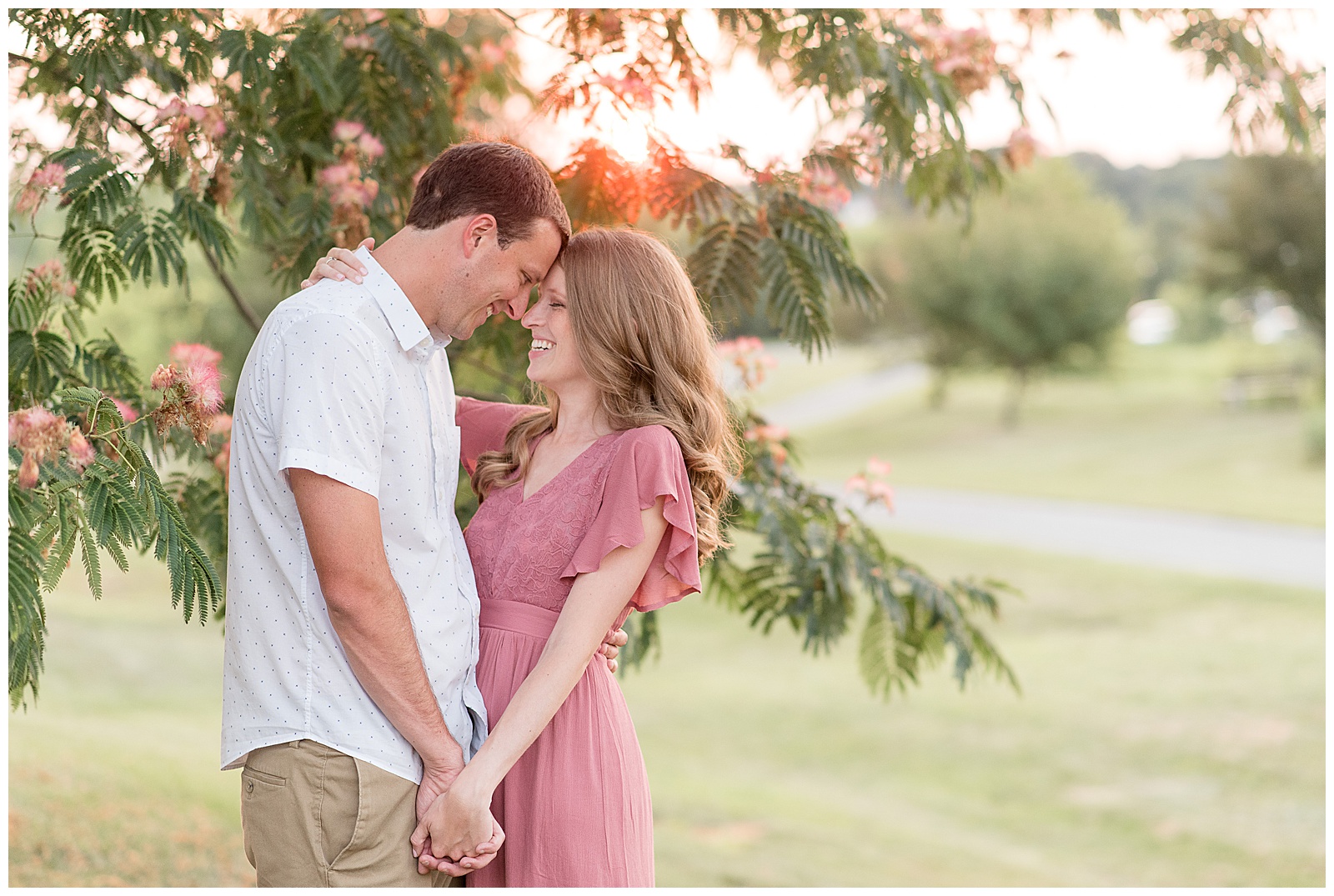 engaged couple standing under tree resting their foreheads together and smiling as they hold hands at sunset