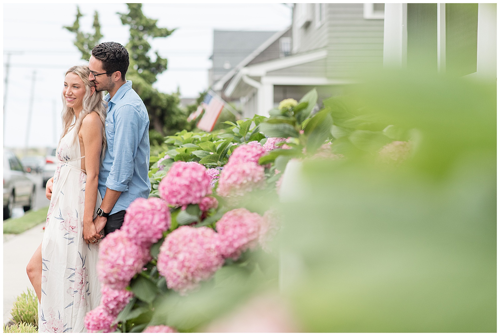 guy standing behind girl and kissing the top of her head along sidewalk with large pink hydrangea bush in beach town by homes