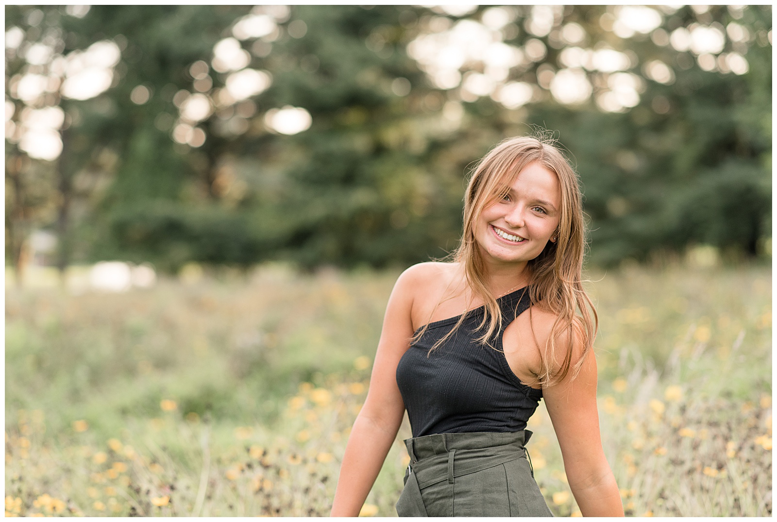 senior girl with left shoulder pressing back smiling at camera wearing trendy black tank top and army green skirt by wild grasses in lancaster pennsylvania