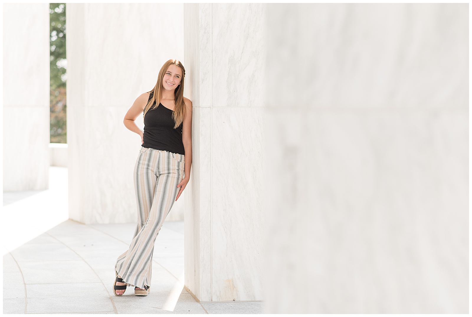 senior girl wearing black tank top and black and white linen pants leaning left shoulder against white wall with ankles crossed at founder's hall