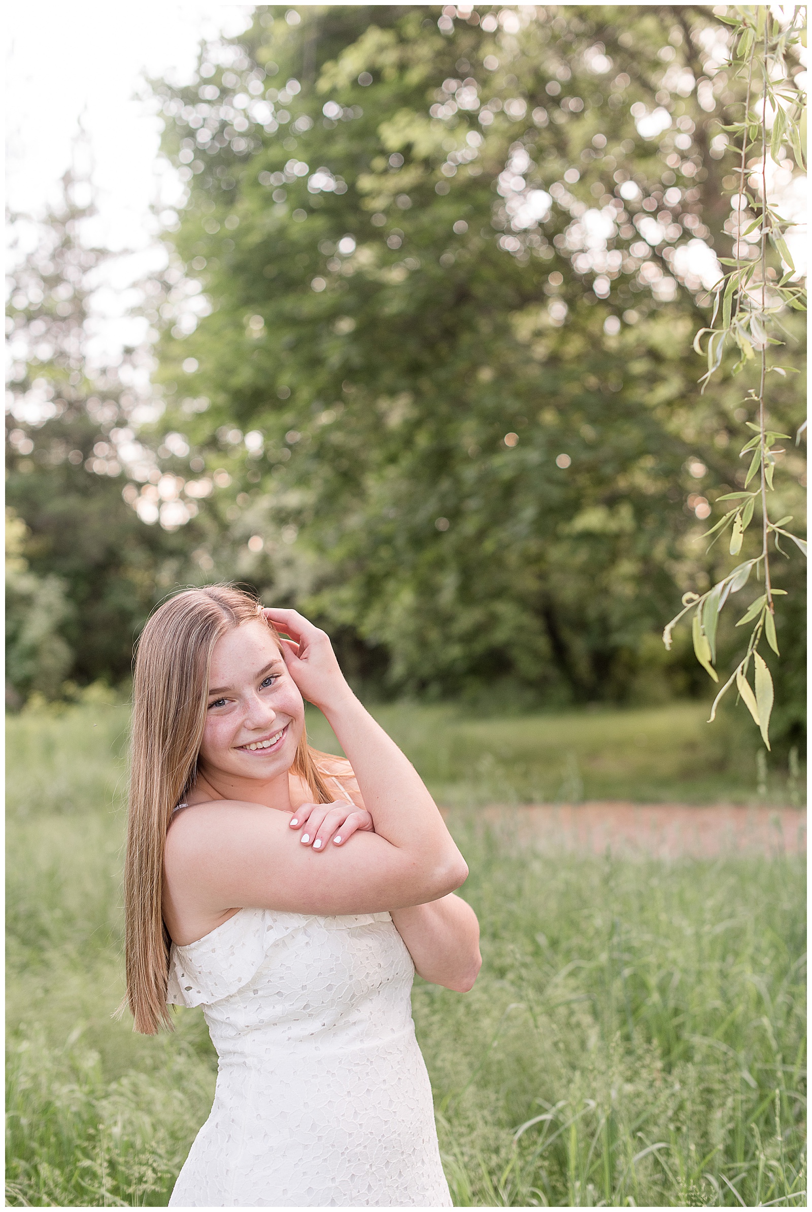 senior girl wearing strapless white summer dress in grass field tucking her hair behind her left ear with her right hand smiling in southeastern pennsylvania