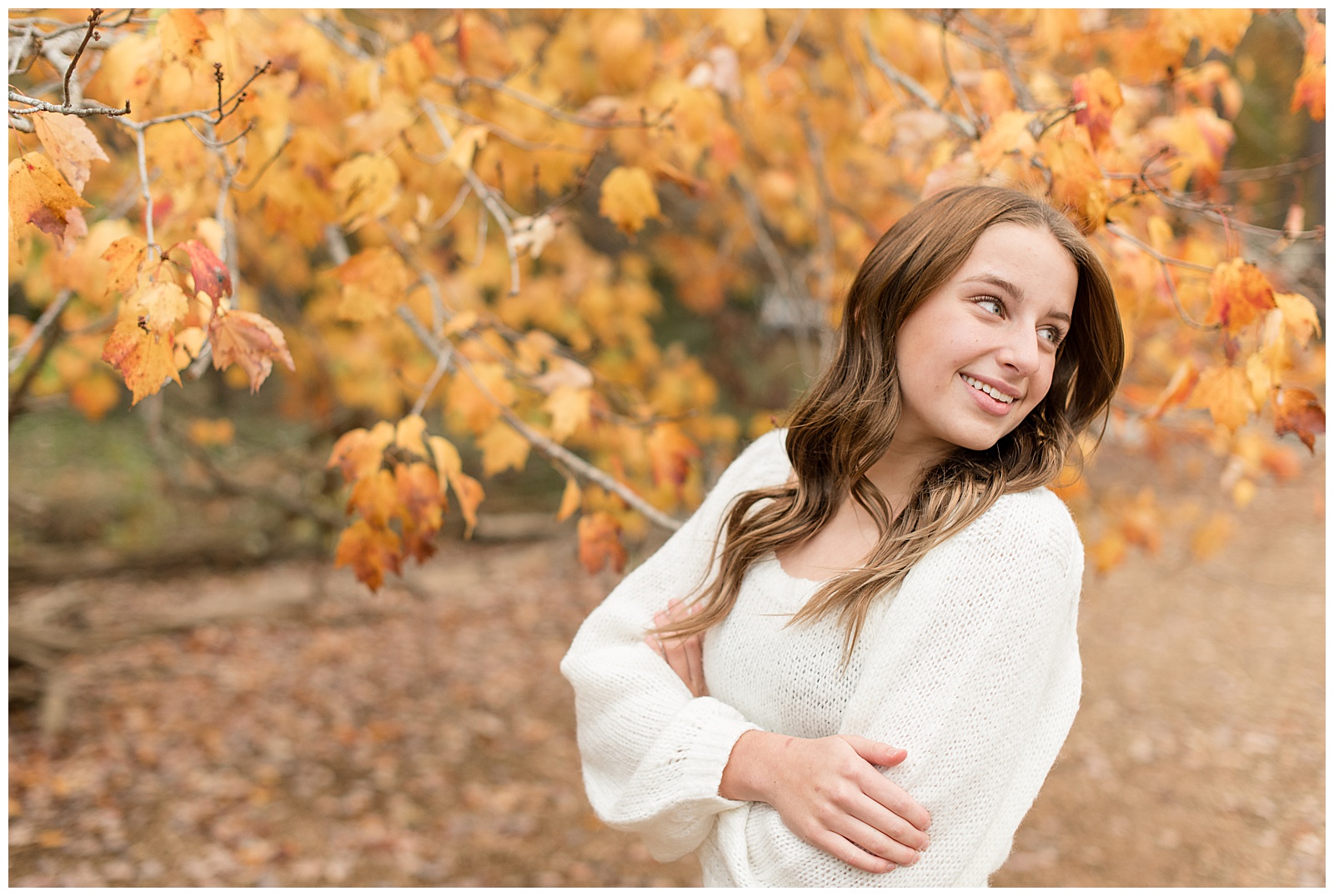 senior girl with arms folded looking back over left shoulder smiling wearing white dress by bright yellow fall tree in southeastern pennsylvania