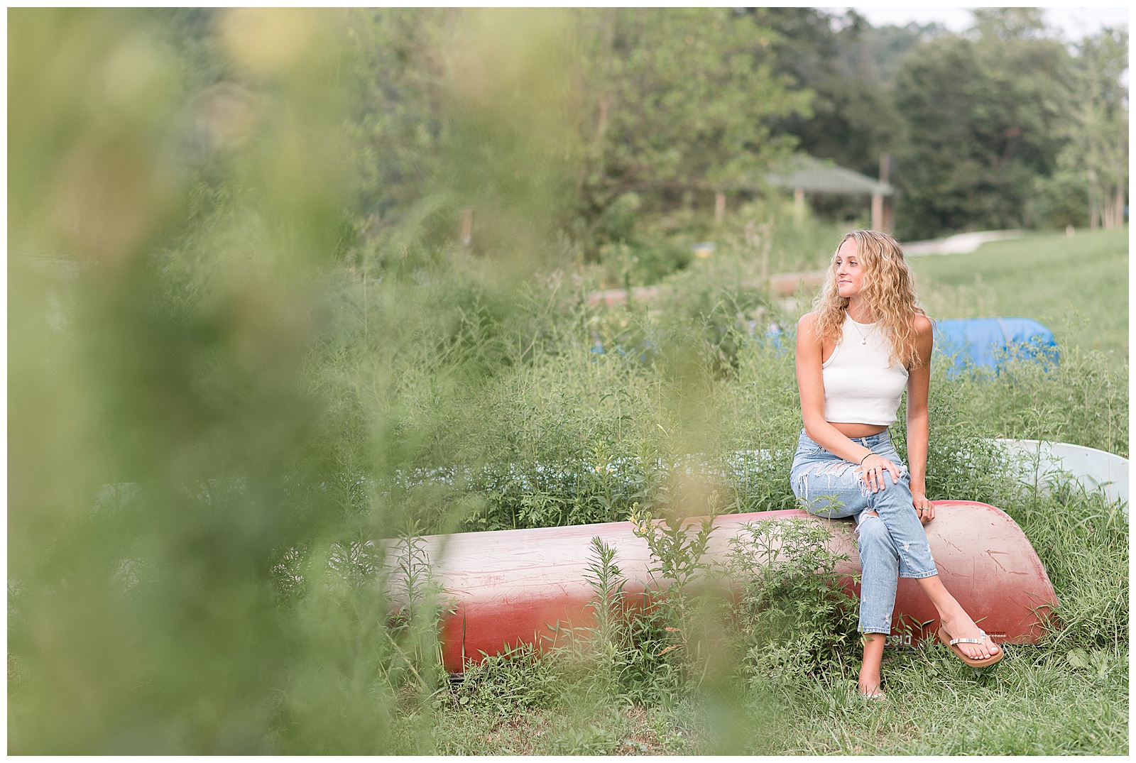 senior girl sitting atop upside down red canoe looking over her right shoulder with her legs crossed at speedwell forge lake in lancaster pennsylvania