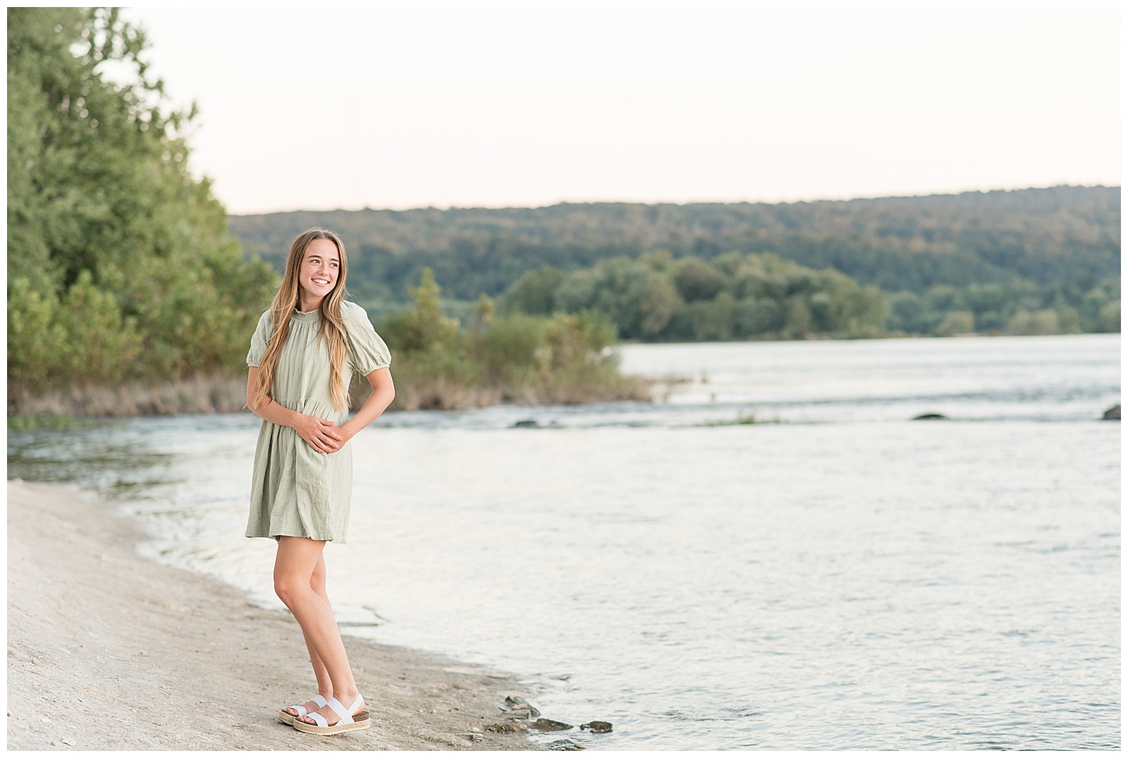 senior girl wearing light green summery dress with both hands on her left hip as she looks towards left shoulder at susquehanna river near white cliffs of canoy