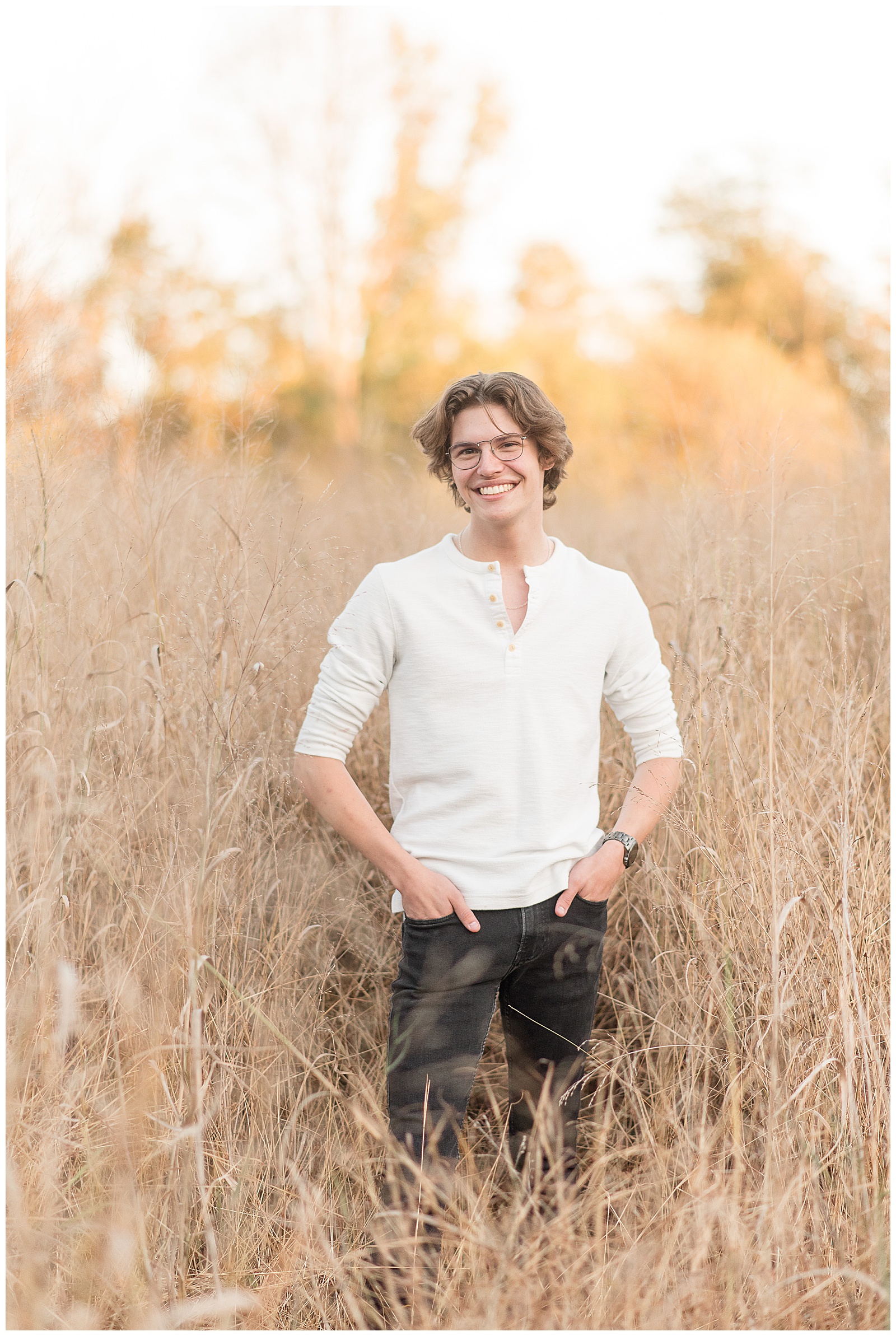 senior guy wearing white shirt and black jeans with his hands in his pockets standing in tall dried wild grasses at sunset in lancaster pennsylvania