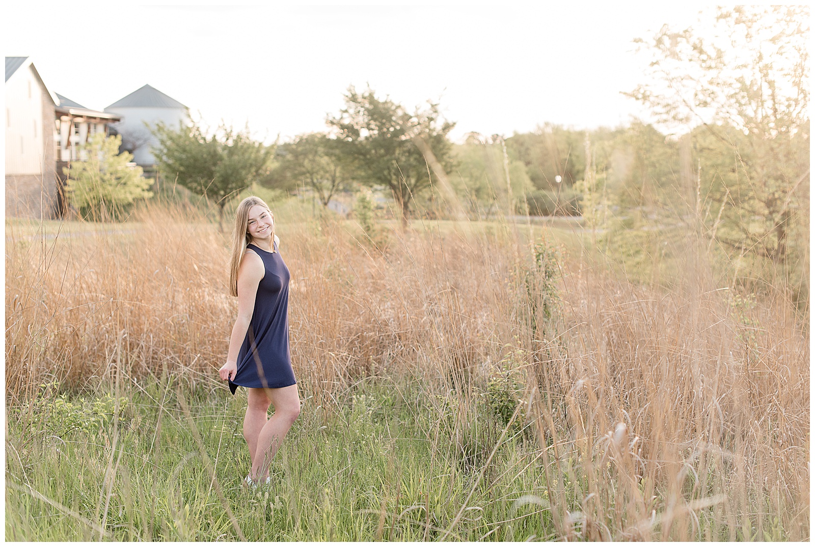 senior girl with right shoulder towards camera standing among field of wild grasses wearing navy blue dress smiling as the sun sets at overlook park
