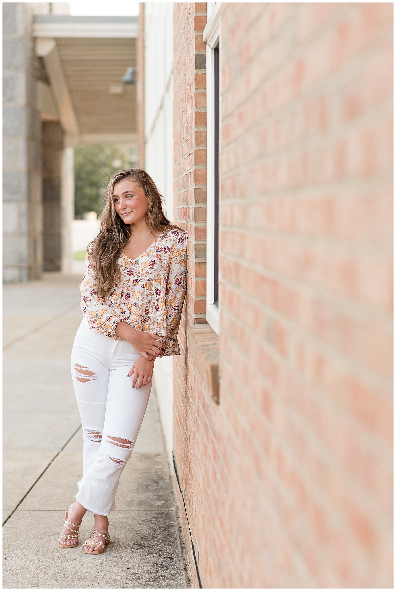 senior girl wearing floral top and ripped white jeans leaning left shoulder against brick building and smiling as she looks towards her right at founder's hall